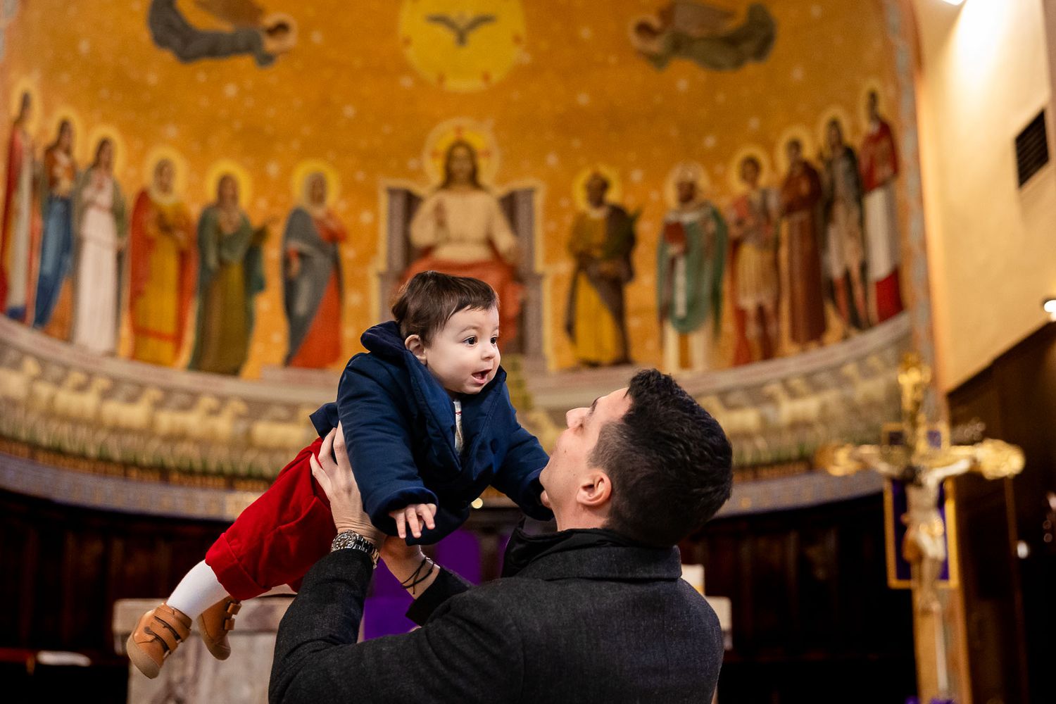 Man lifting a baby in a church with a golden mosaic mural of Jesus and saints in the apse behind them.