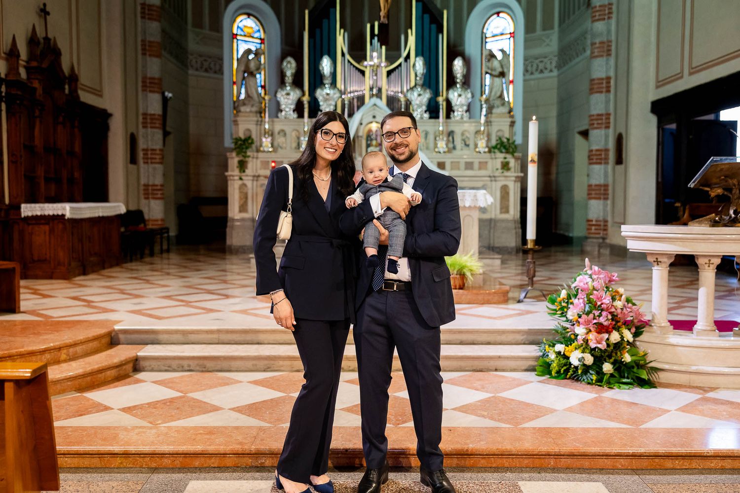 Family posing inside a church during a baptism ceremony, with ornate altar and floral arrangements in the background.