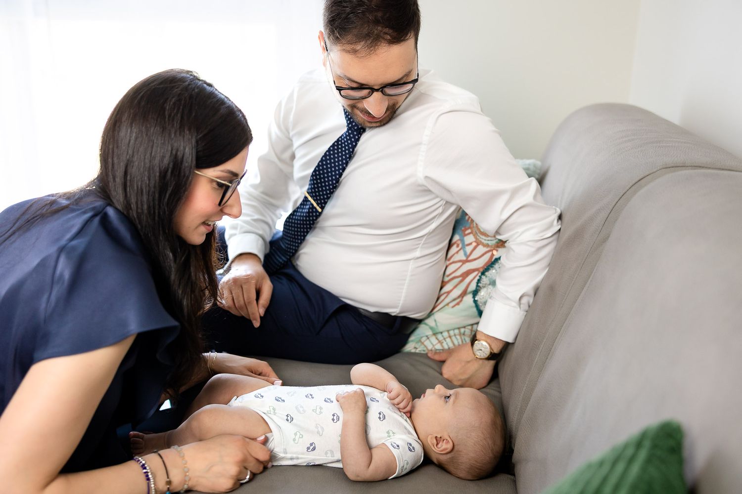 Parents smiling and bonding with their newborn baby lying on a gray sofa in a bright living room.