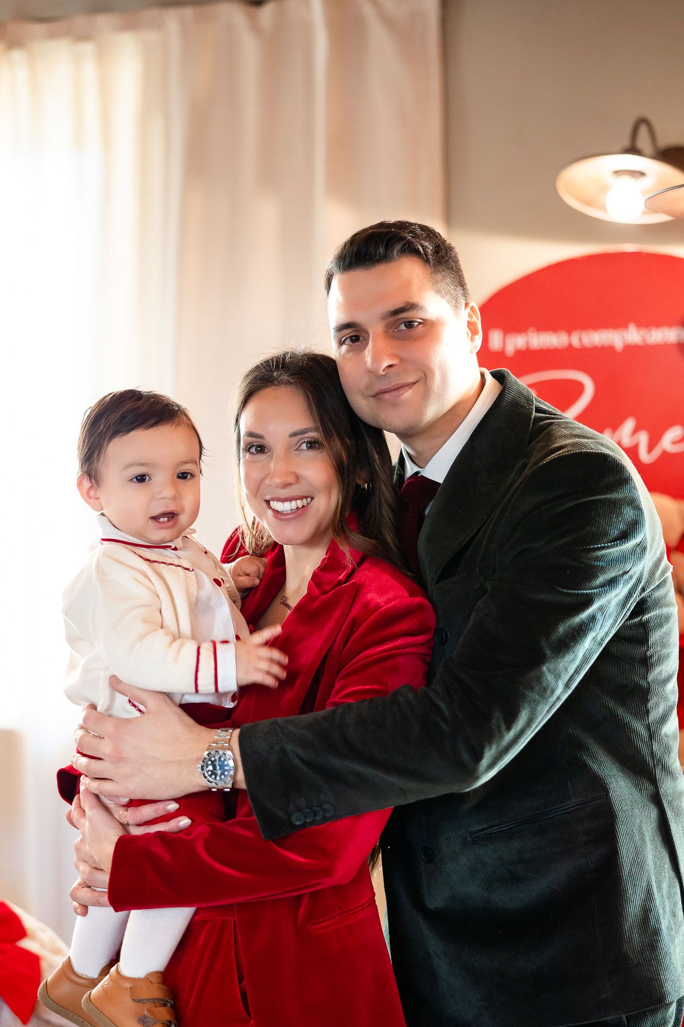 A smiling couple in red and green outfits holds their baby boy at a first birthday celebration.
