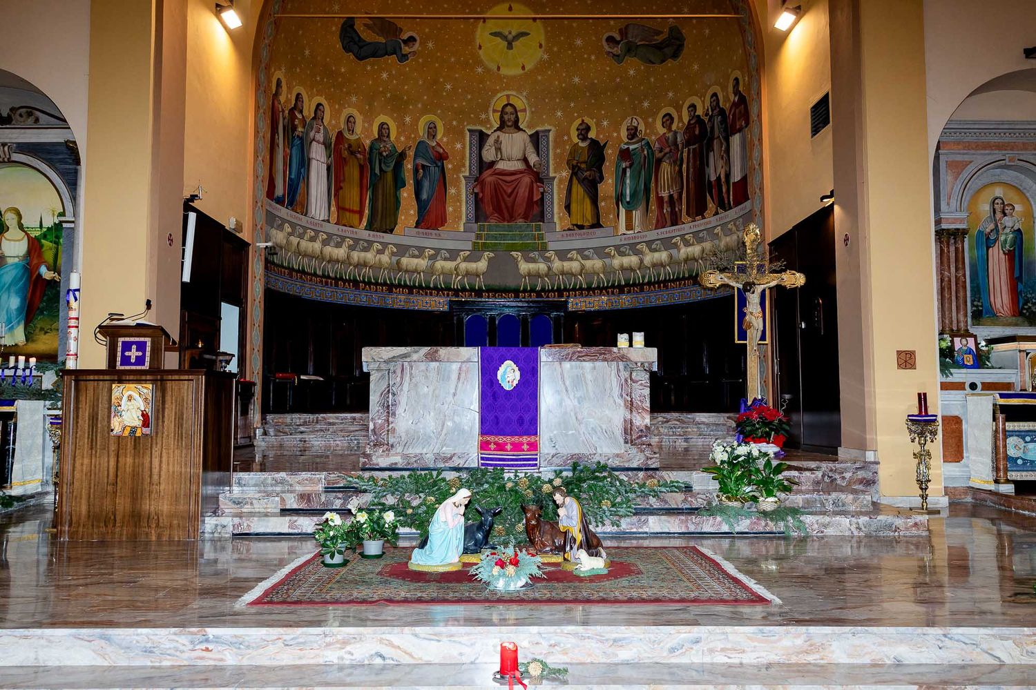 Catholic church interior with ornate mosaic apse, altar adorned with purple cloth, nativity scene, and floral arrangements.