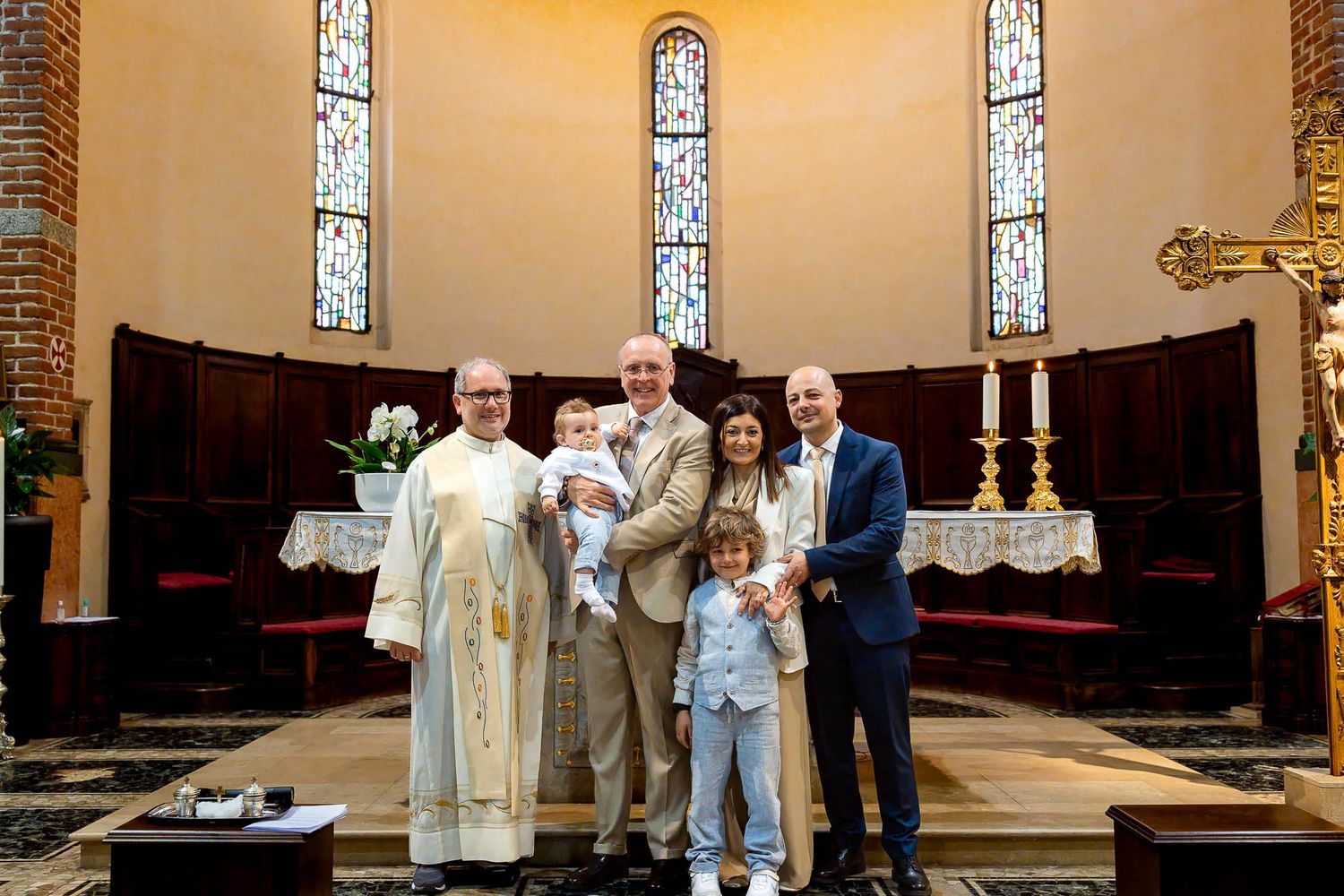 Family posing with priest at church altar during baptism ceremony, featuring stained glass windows.