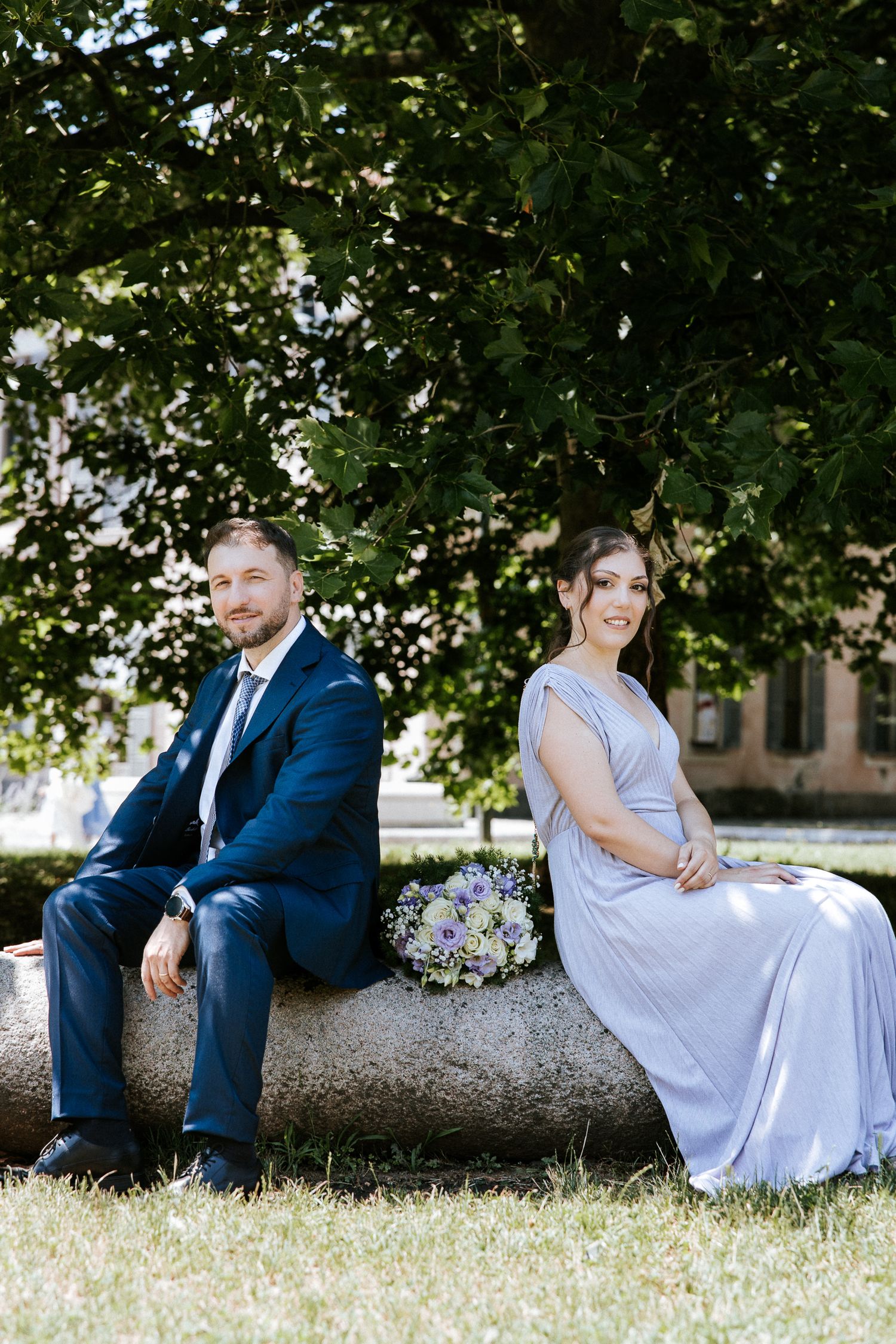 A couple sits back-to-back on a stone wall outdoors, with green trees and a purple bouquet in the background.