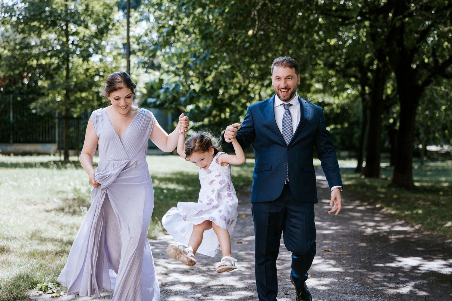 Family walking together on a tree-lined path, with a young child jumping joyfully between two adults in formal attire.