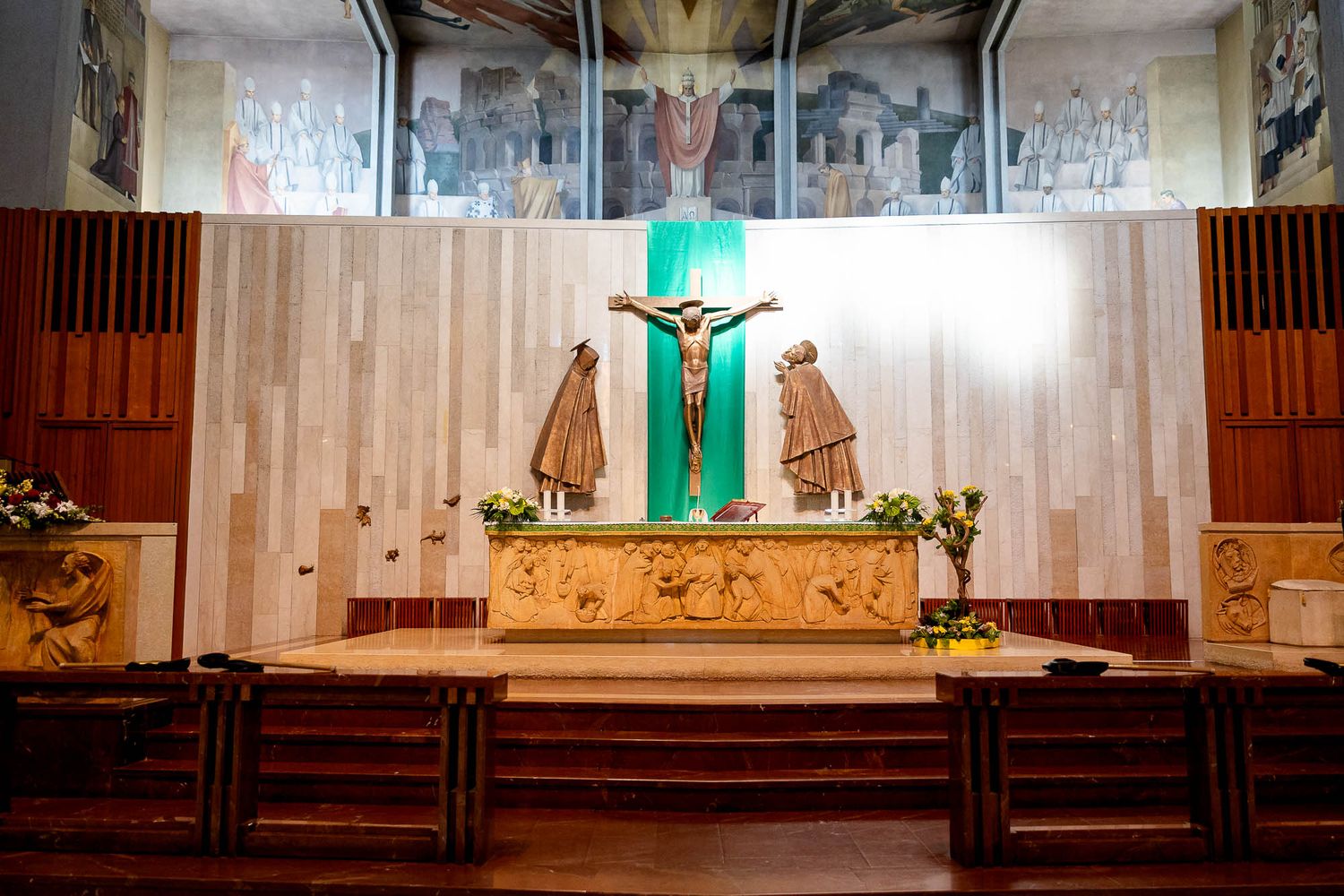 Catholic church altar with crucifix, bronze saints, and decorative mosaic panels in a modern sanctuary.