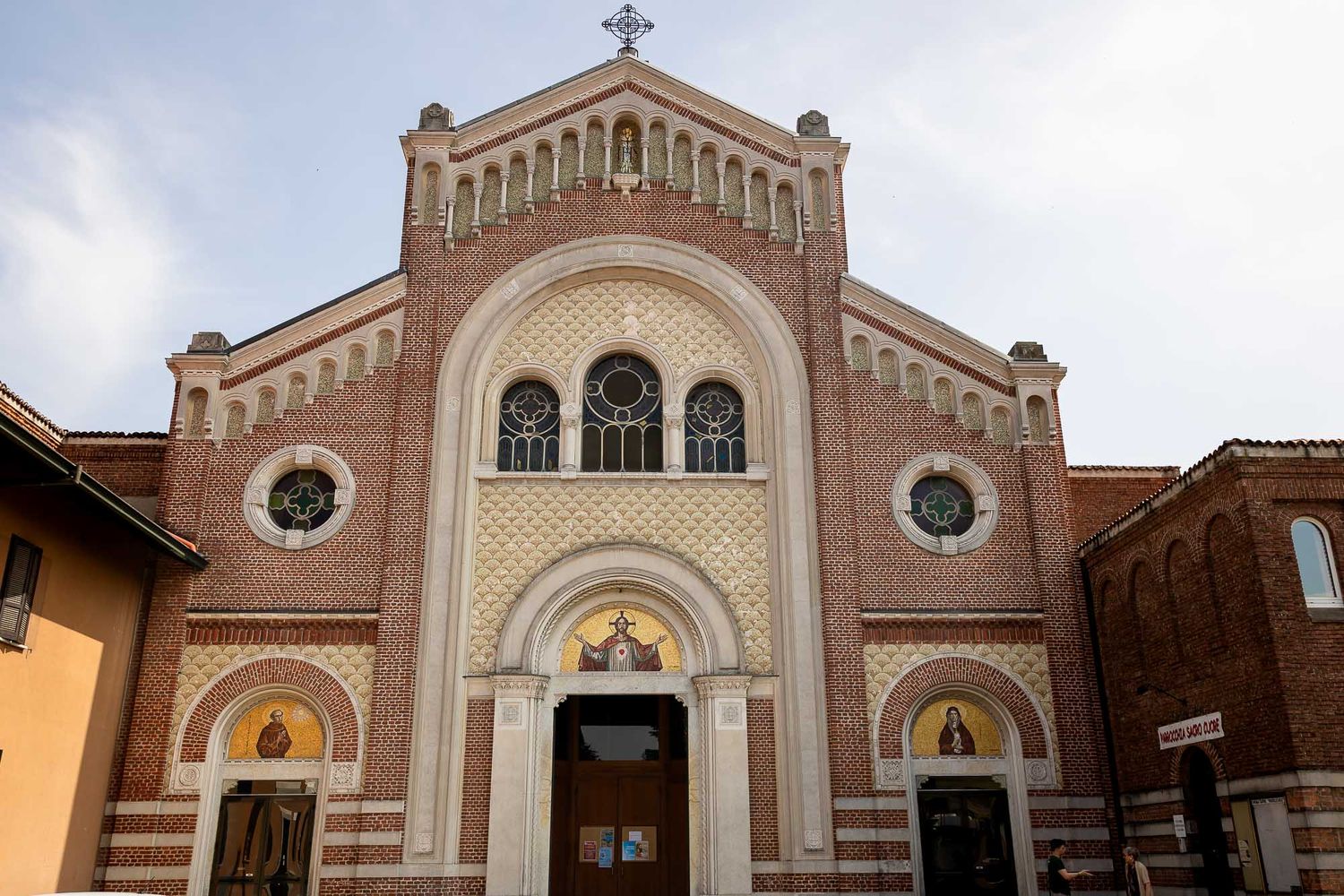 Ornate brick Catholic church facade with arched windows, mosaic details, and a cross atop the gabled roof.
