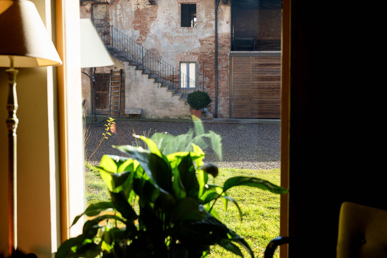 View through a doorway of a sunlit courtyard with green lawn, brick building, outdoor staircase, and lush indoor plants in foreground.