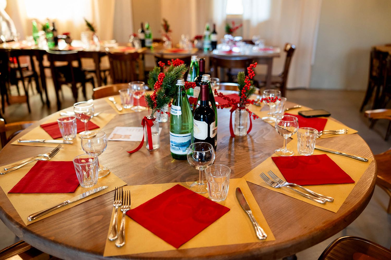 Round dining table set with red napkins, gold placemats, wine glasses, and bottles as centerpiece in elegant venue.