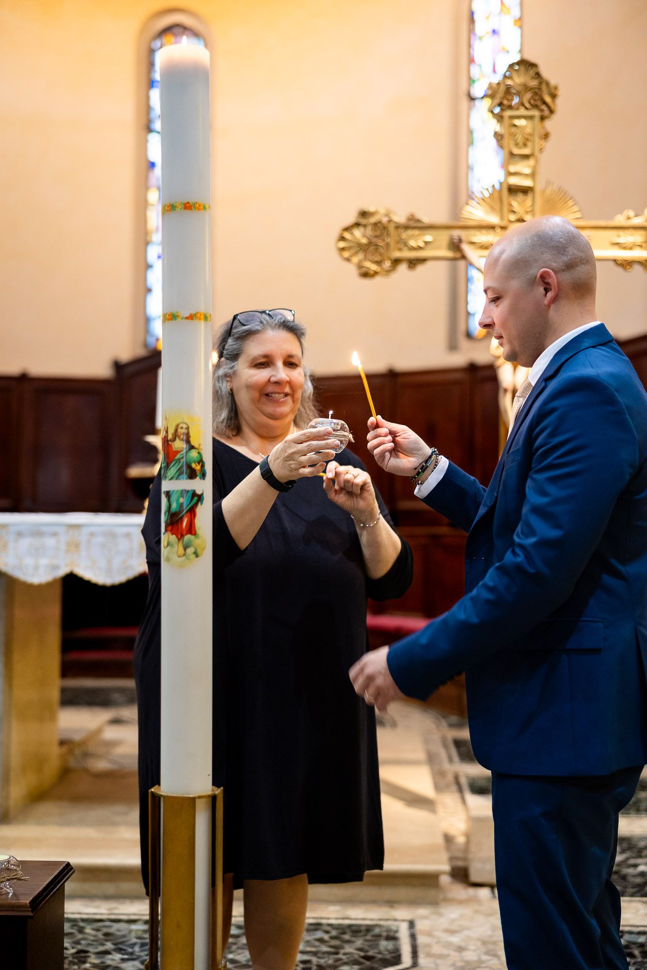 Woman in black dress and man in blue suit lighting candles near a large paschal candle inside a church.