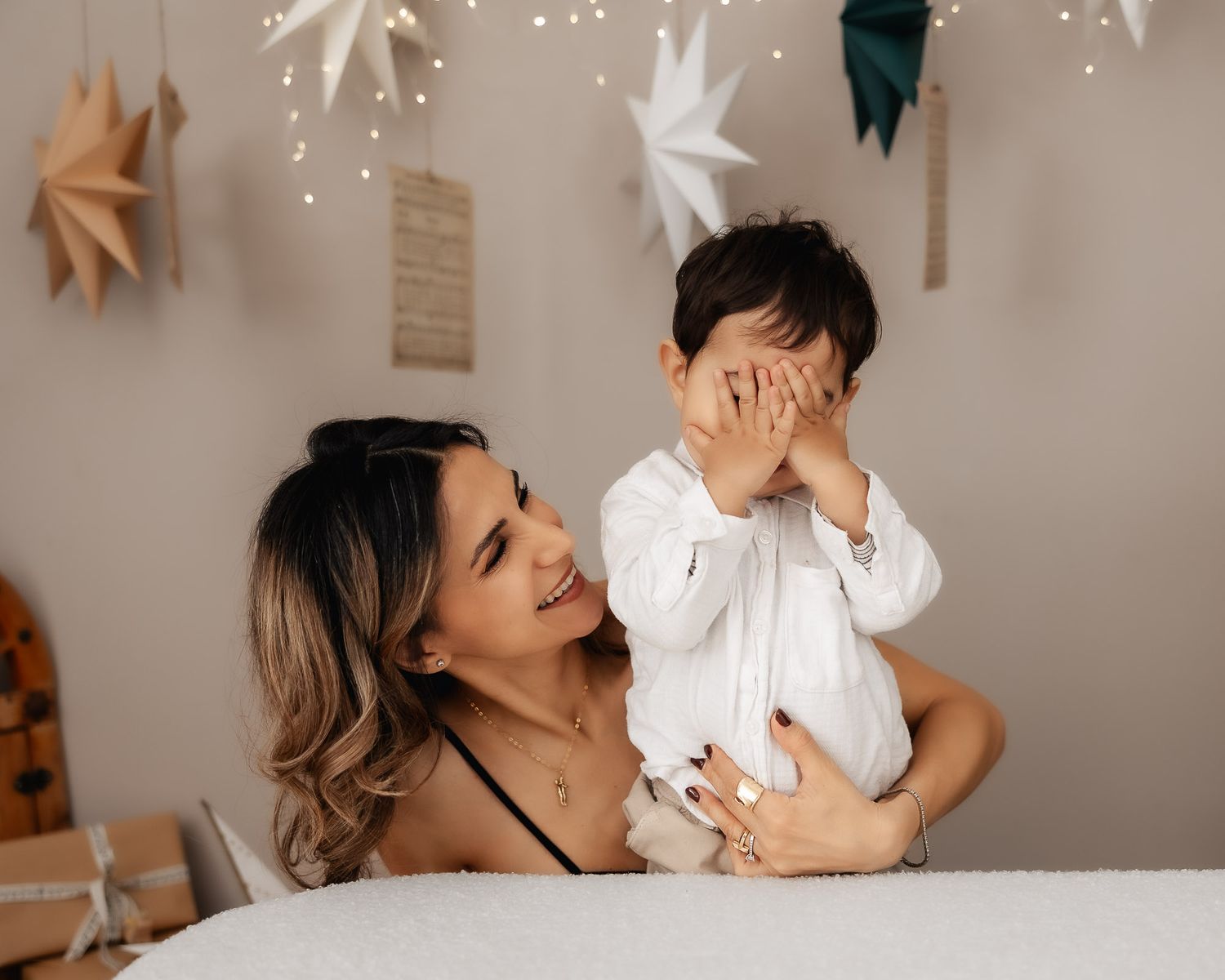 A smiling mother holds her baby boy who covers his eyes with his hands, surrounded by festive star decorations.