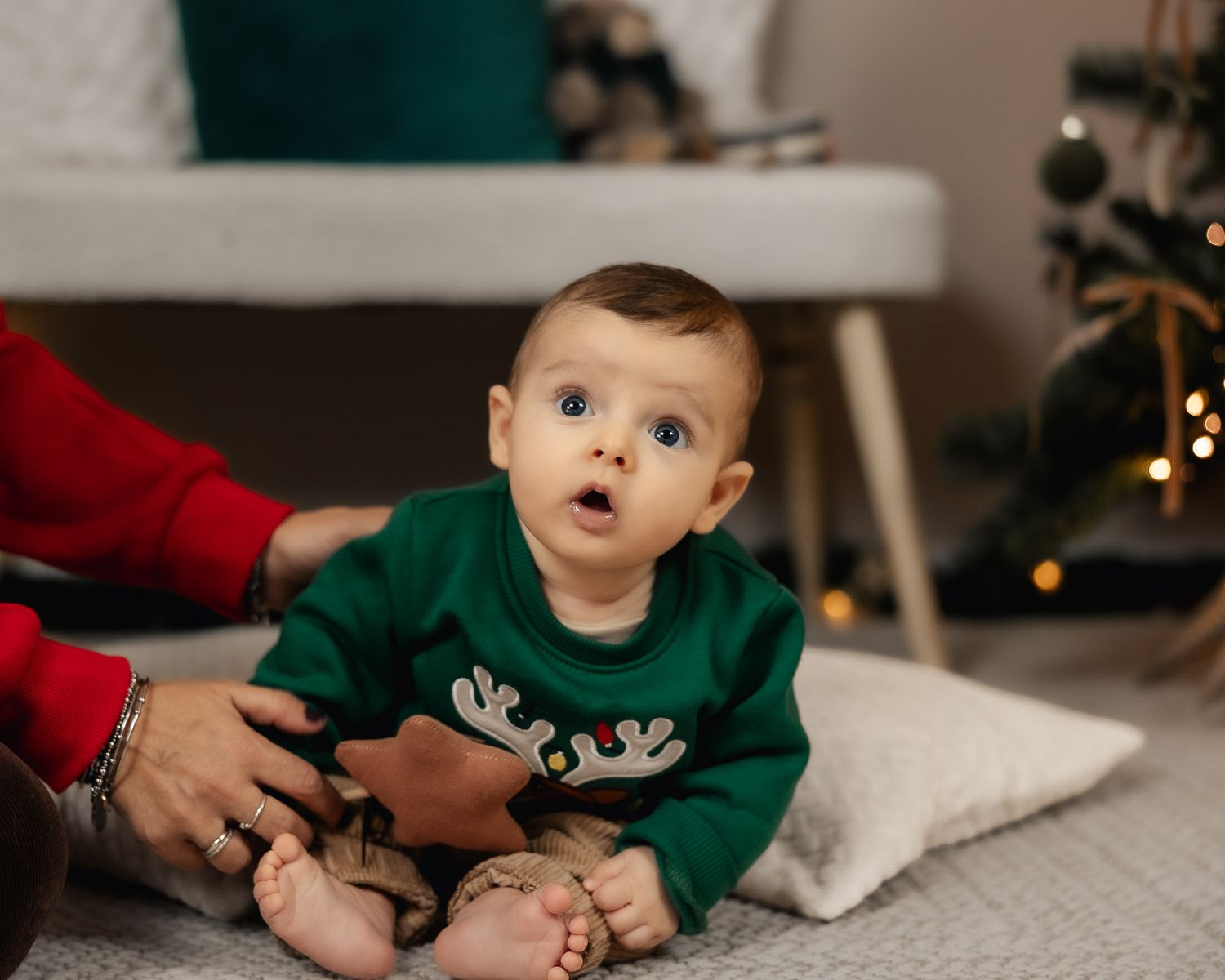 A wide-eyed baby in a green reindeer Christmas sweater sits on a white rug near a decorated holiday tree.