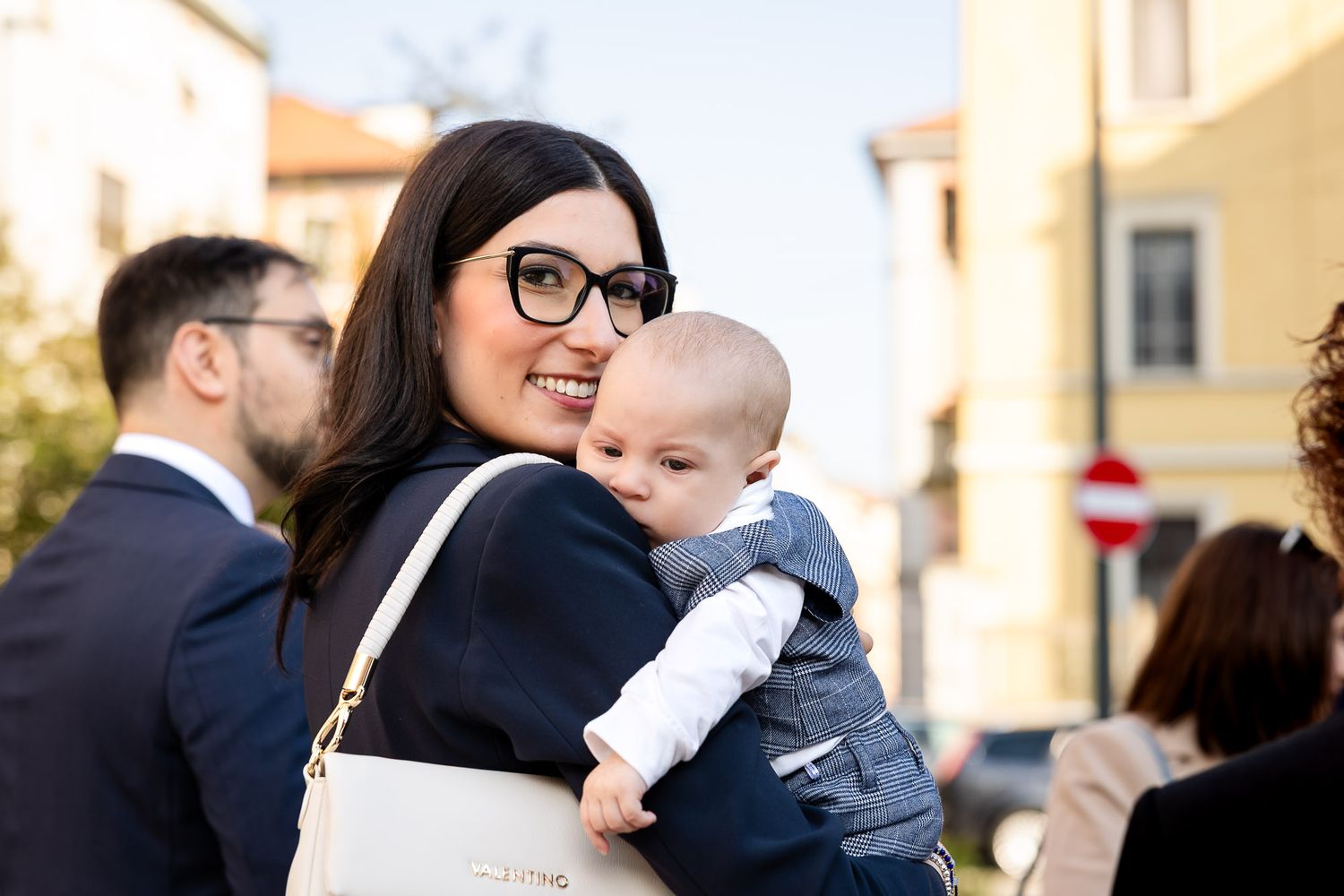 Smiling woman with glasses holds a baby in a suit outdoors at a sunny urban event.