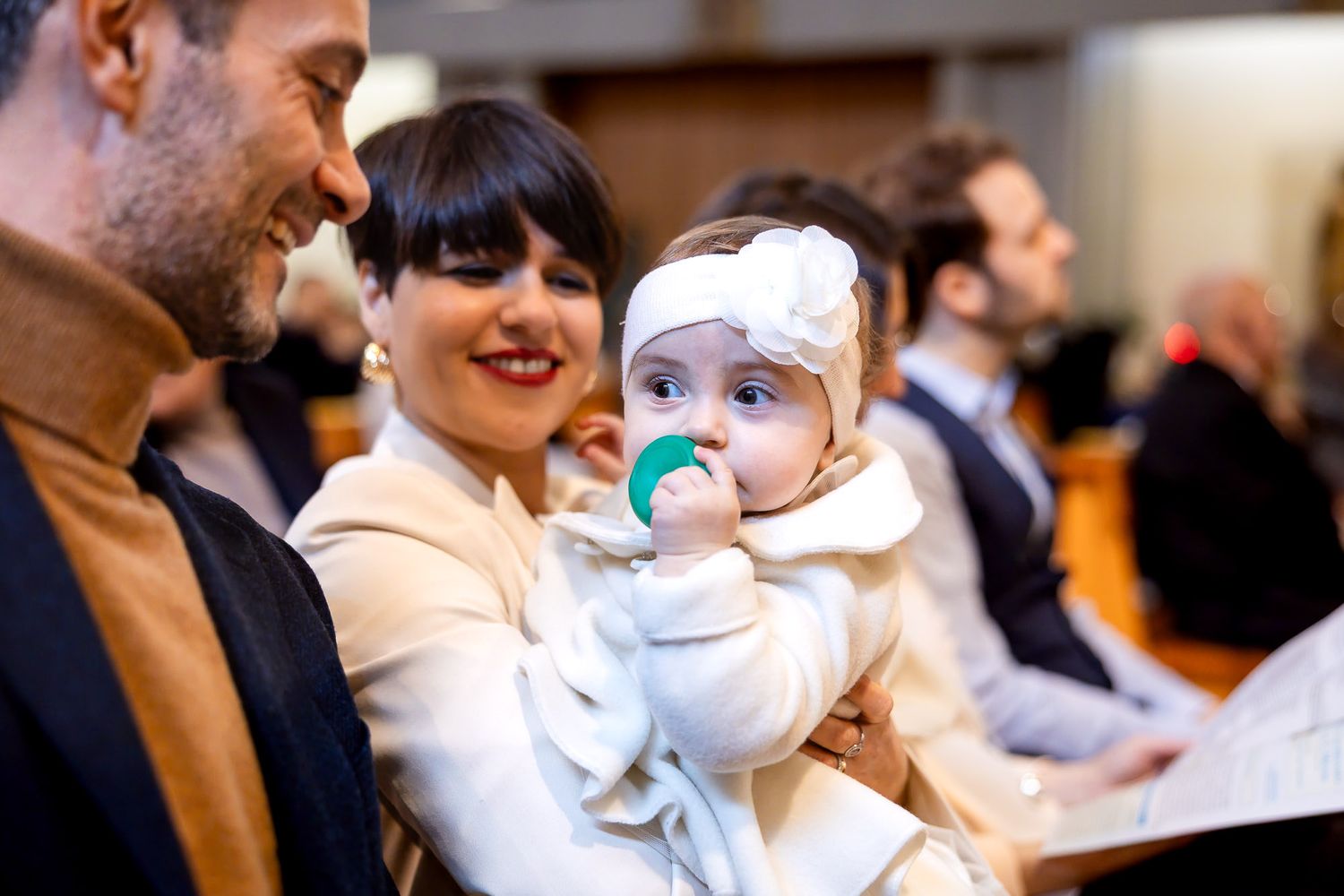 A smiling mother holds her baby girl in a white outfit with a bow headband and blue pacifier at a church event.