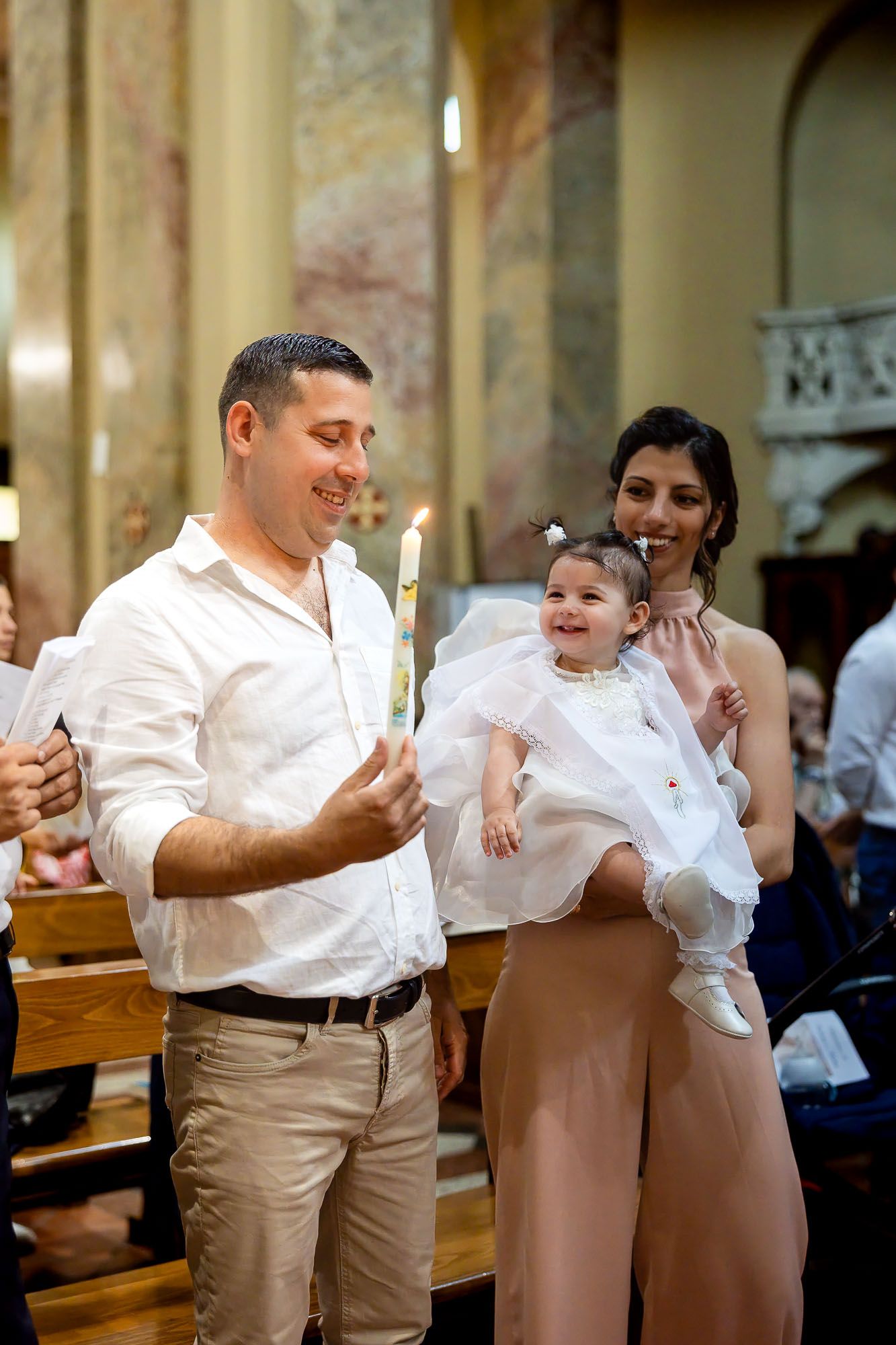 Family celebrating a baby baptism in church, parents holding infant in white christening gown with lit candle.