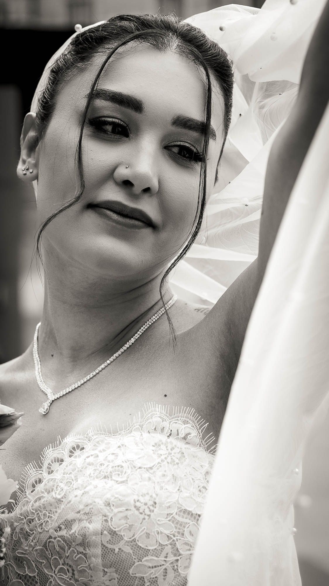 Black and white portrait of a bride in lace wedding dress and veil, wearing a delicate necklace, looking down gracefully.