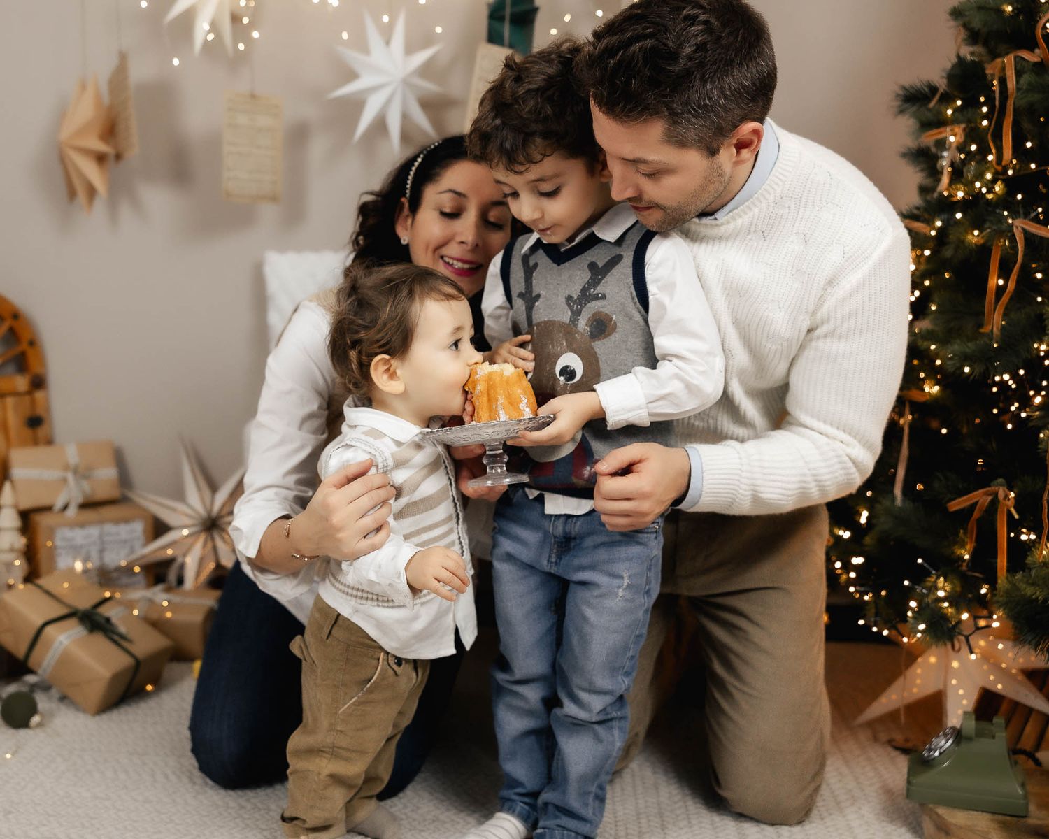 Family of four celebrating Christmas near a decorated tree, with parents and two young children sharing a festive gift moment.