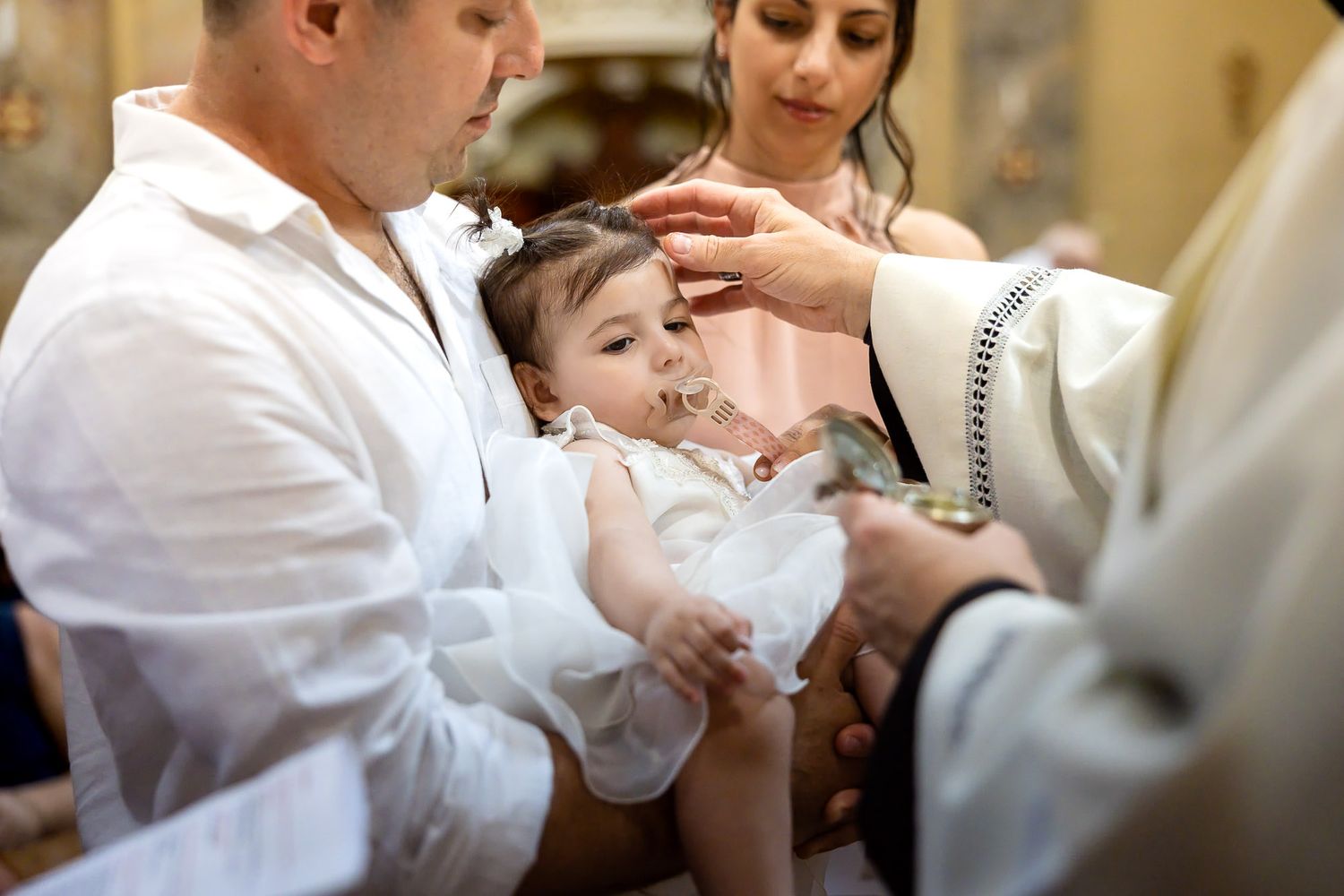 Priest performing Catholic baptism ceremony on infant in white dress, held by father with mother present.