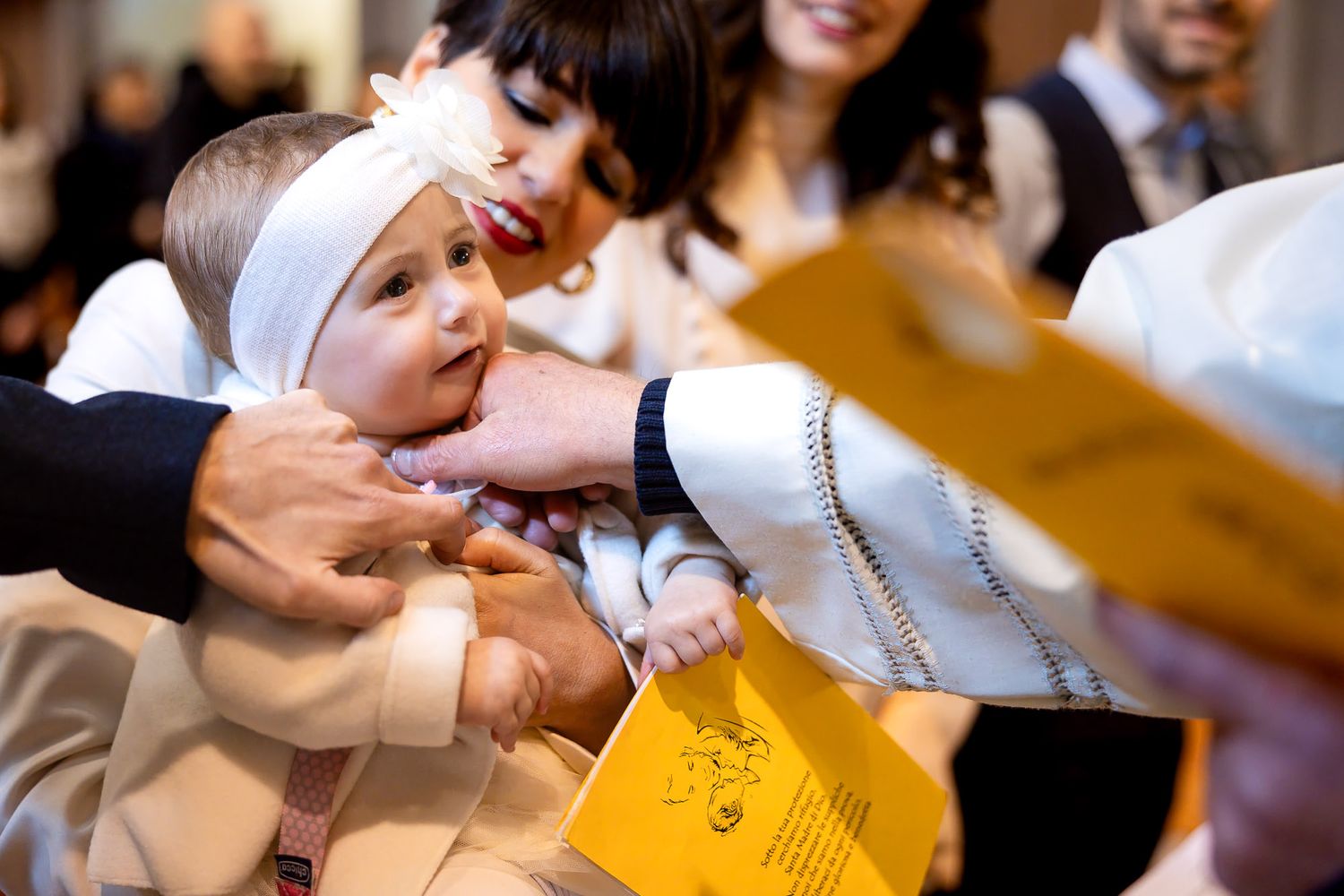 Baby in white bonnet held during baptism ceremony with priest and family members present.