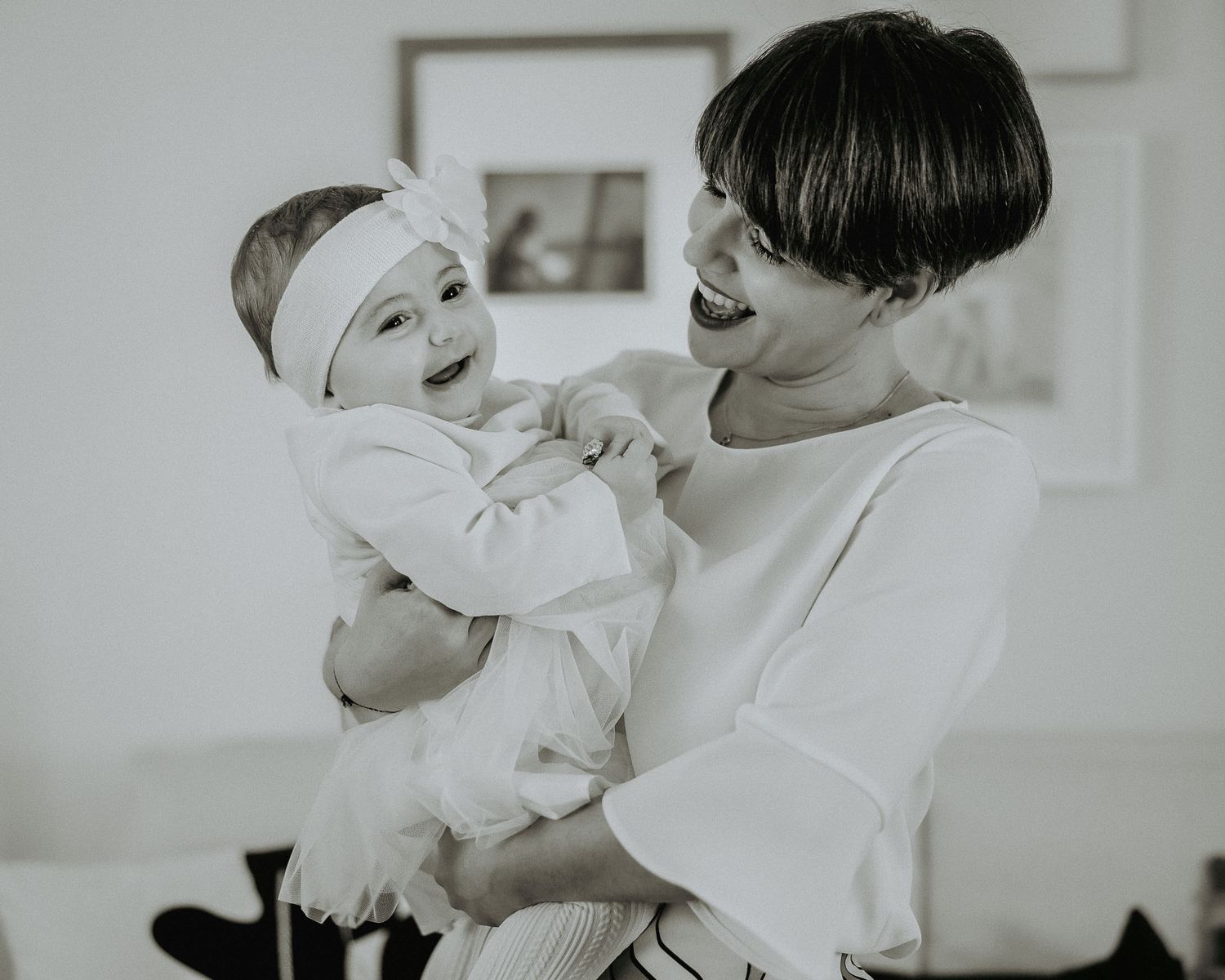 Black and white photo of a laughing mother holding her smiling baby girl with a white bow headband.