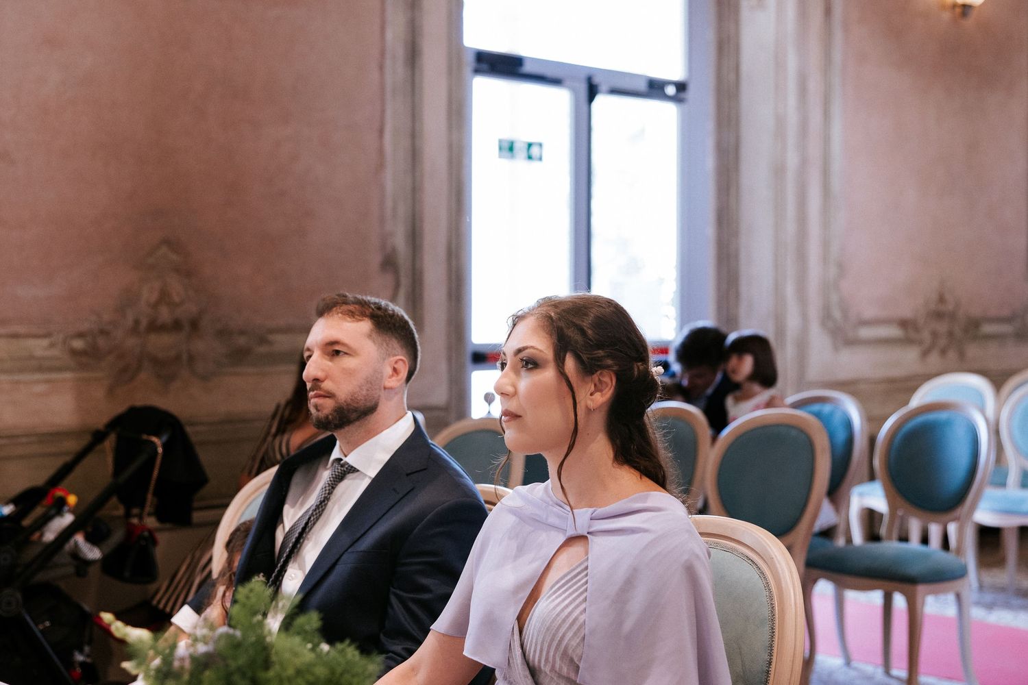 Wedding guests seated at elegant reception tables in an ornate ballroom with crystal chandeliers overhead.