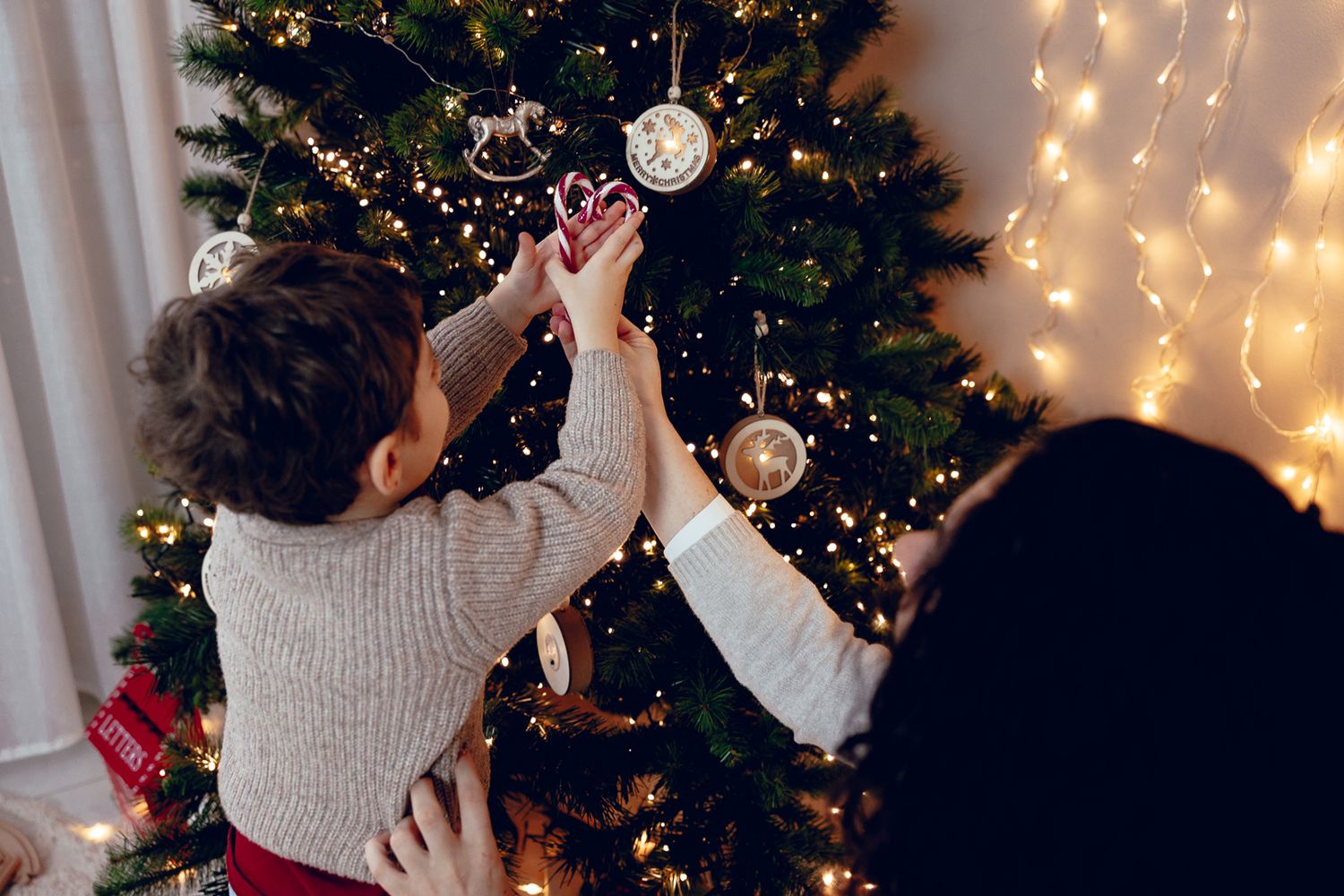 A person decorates a Christmas tree with ornaments and lights while wearing a cozy sweater.