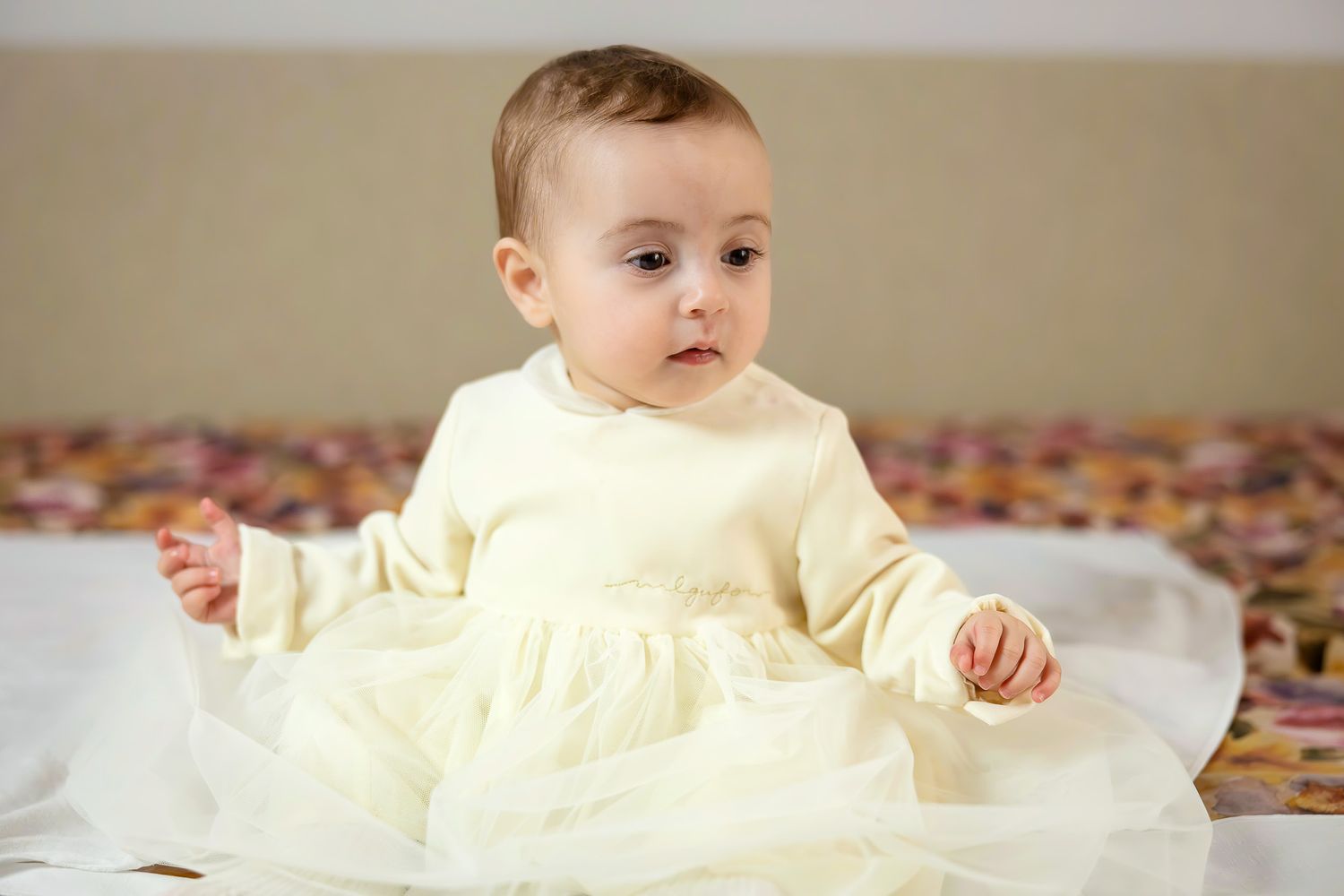 Adorable baby girl in a white tulle dress sitting on a bed, looking curiously to the side.