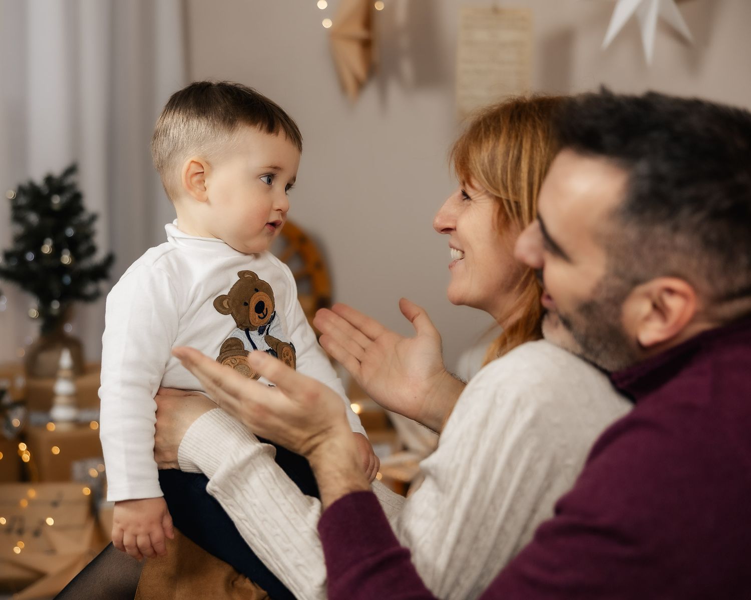 A happy family with parents holding their baby boy near Christmas decorations and warm fairy lights.