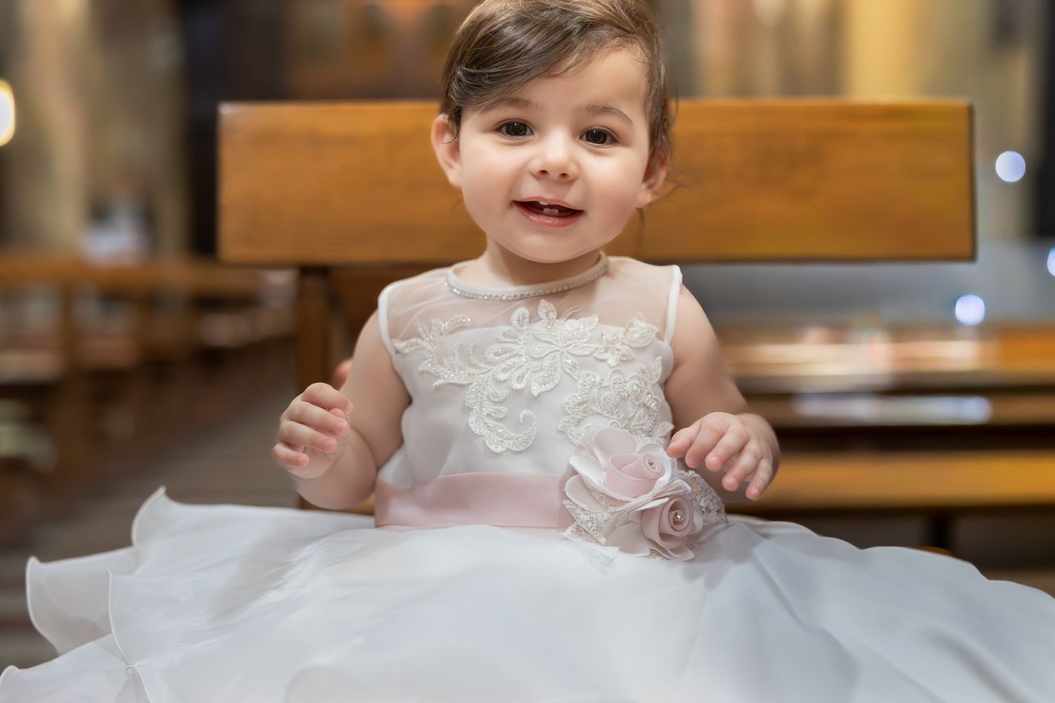 An adorable baby girl smiling in a white lace christening gown inside a church.