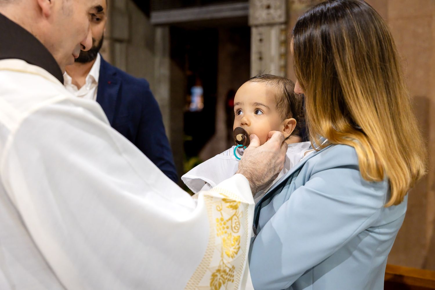 Priest performing a baptism ceremony on a baby with a pacifier held by a woman in a light blue jacket.