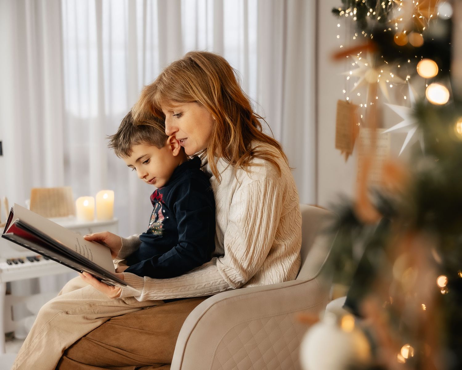 A mother and child read a book together in a cozy armchair beside a decorated Christmas tree with warm glowing lights.