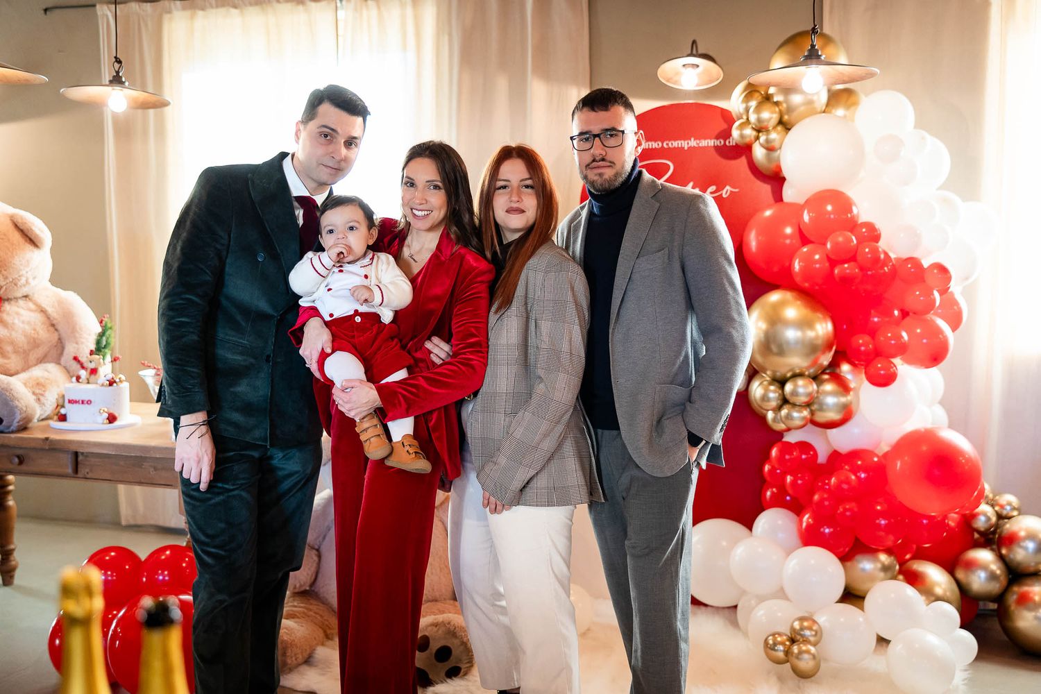 Family posing together at a festive red and white balloon-decorated celebration party indoors.