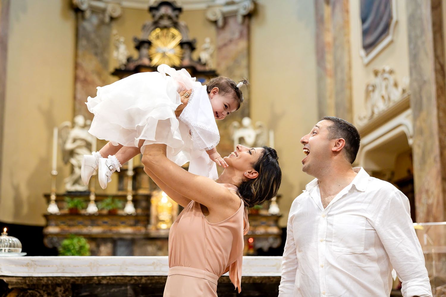 Joyful parents lift their baby in white dress during a christening ceremony in a church.