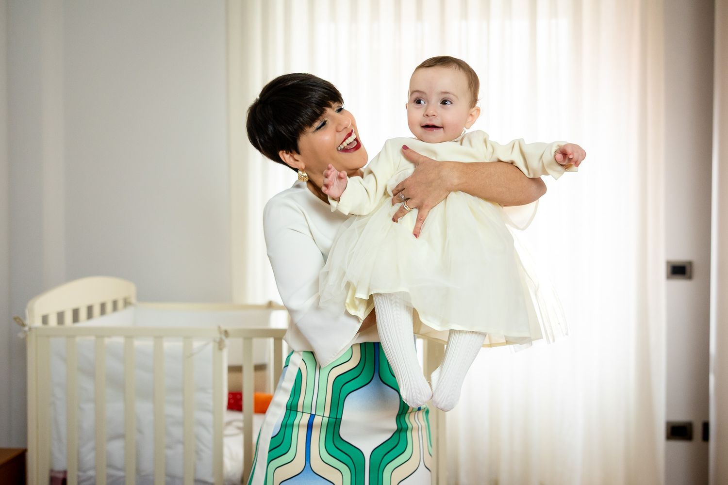 Joyful mother lifting her smiling baby girl in a cream dress near a white crib in a bright nursery.