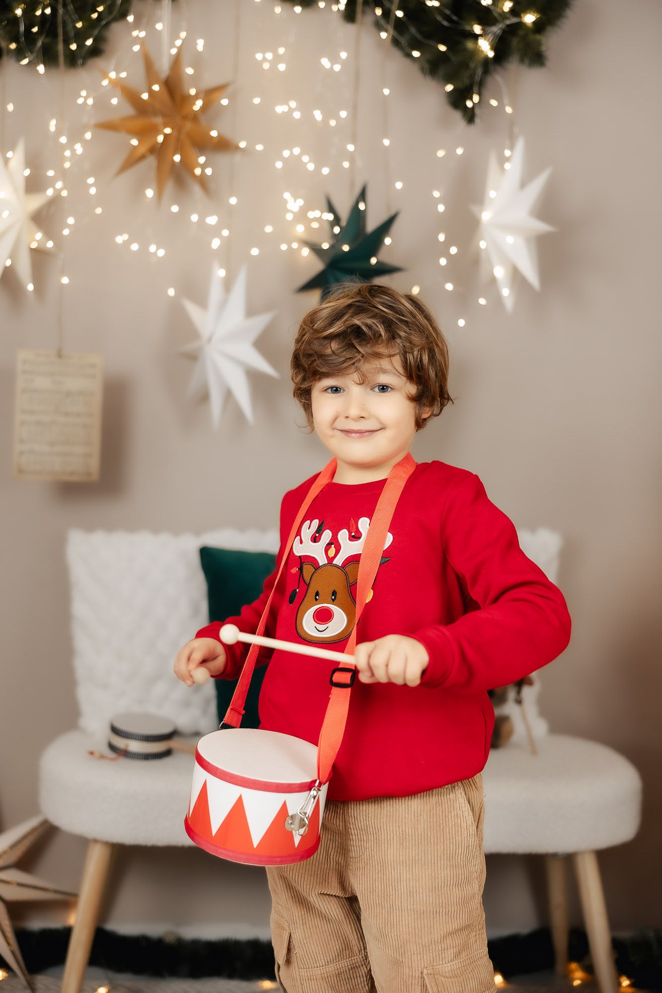 Boy in red reindeer sweater playing toy drum amid Christmas star decorations and fairy lights.