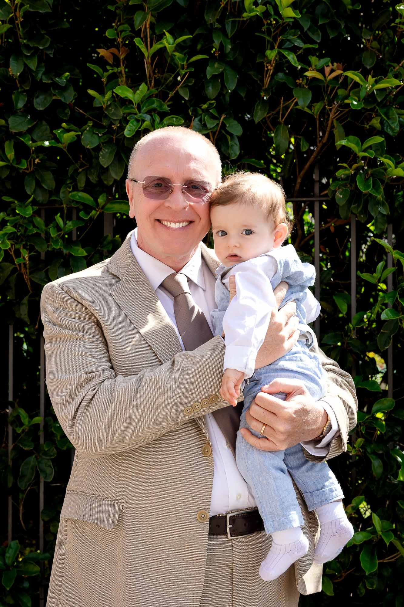 Smiling bald man in tan suit holding a baby in denim outfit outdoors with green foliage background.