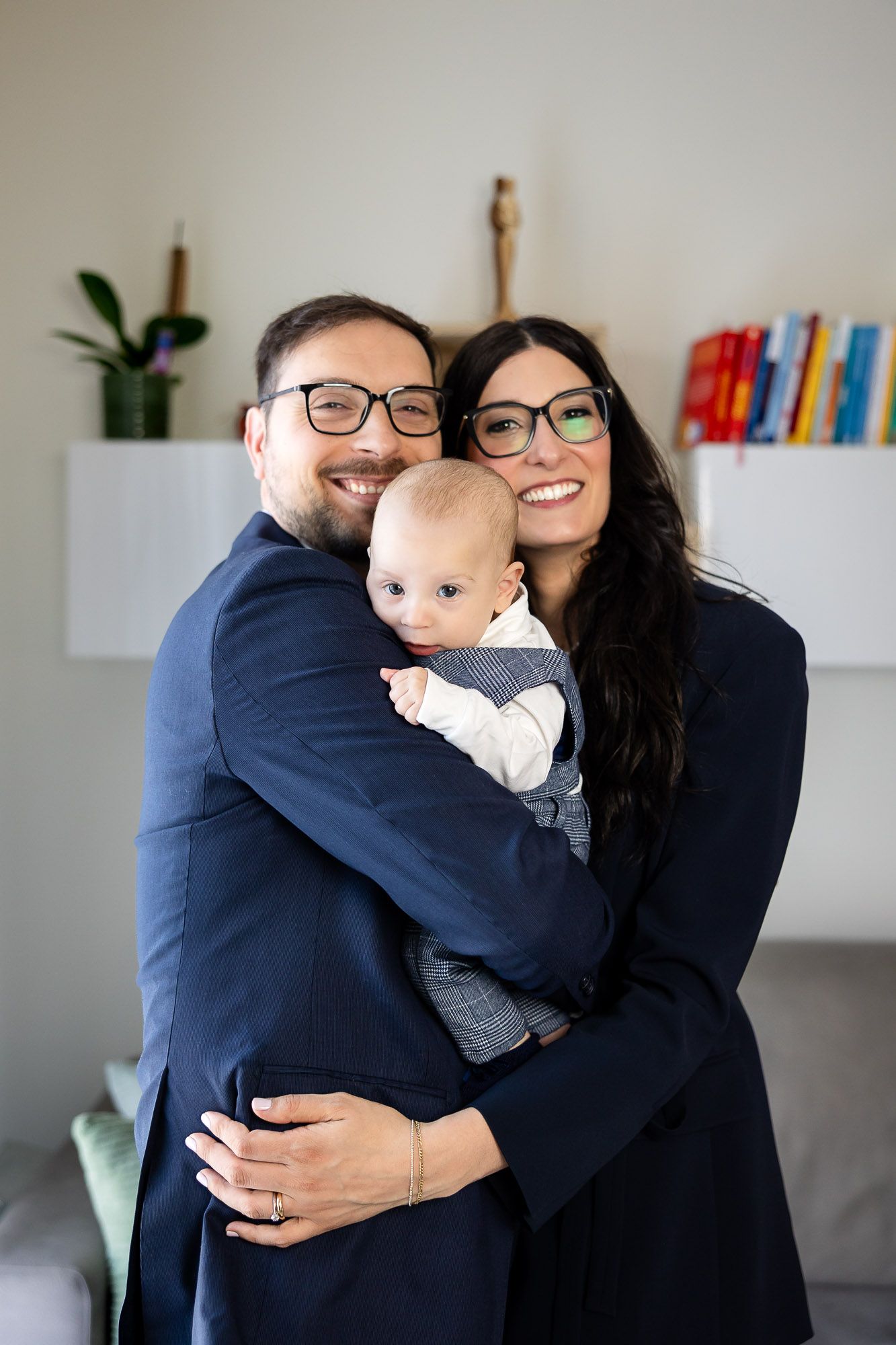 Happy family portrait with parents wearing glasses and navy suits holding their infant baby.