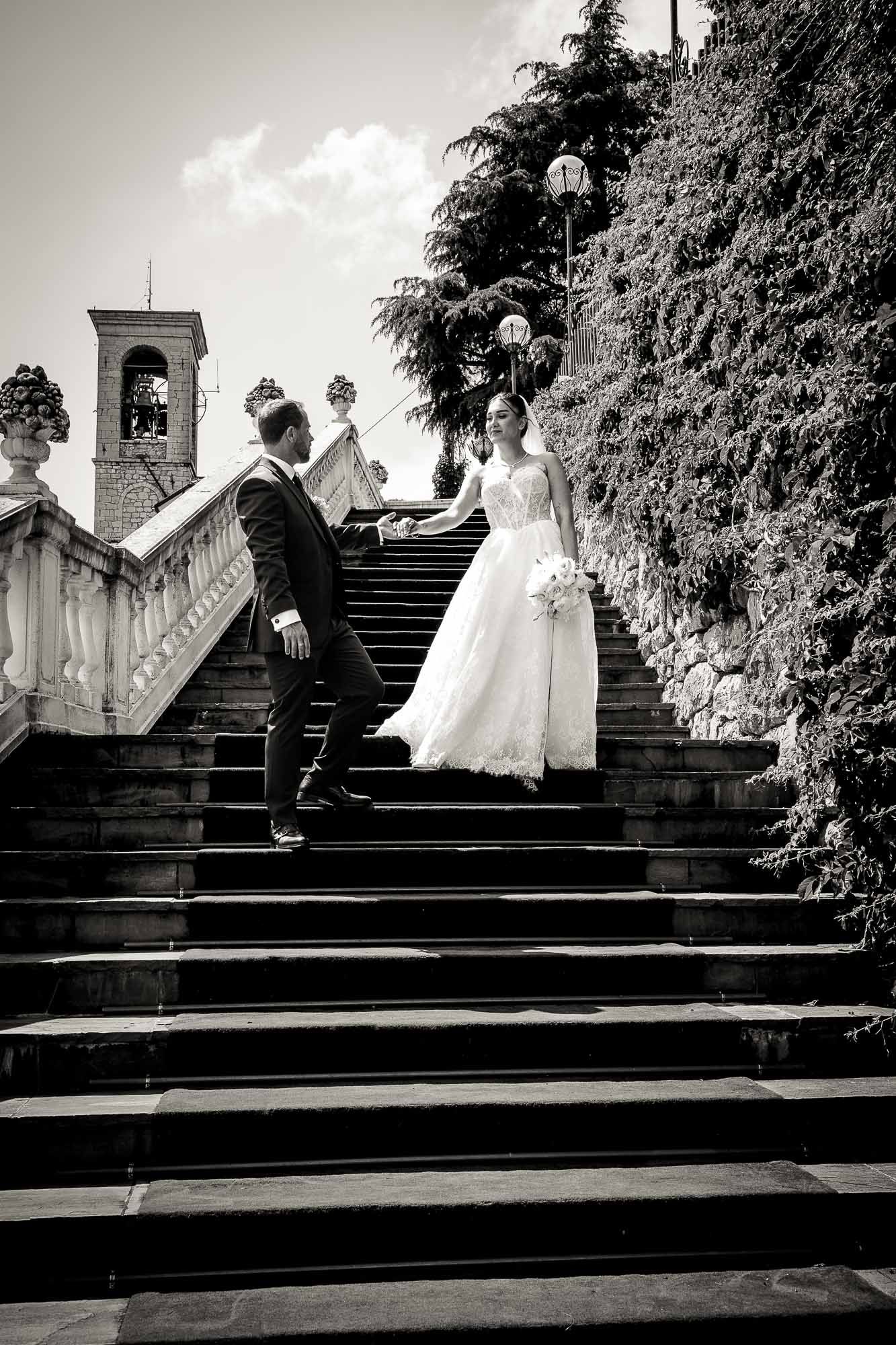 Black and white photo of bride and groom holding hands on elegant stone staircase at outdoor wedding venue.