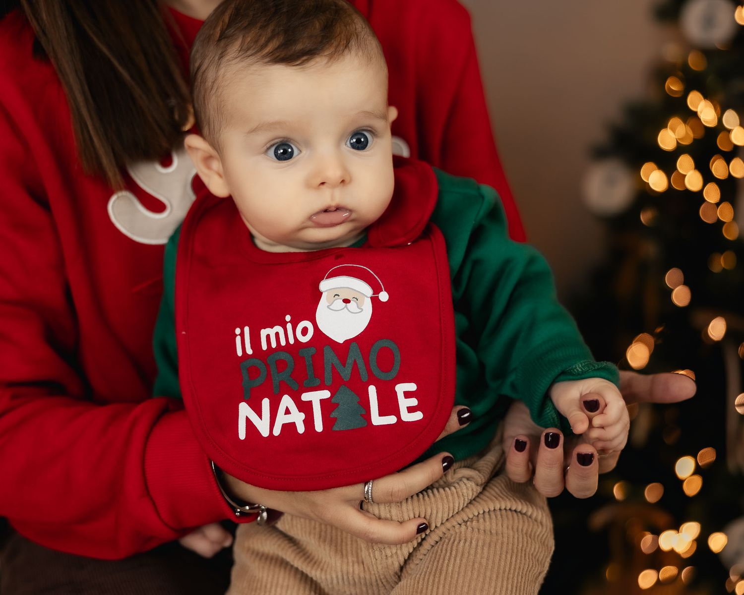 Baby wearing a red bib saying 'il mio primo Natale' held by a person near a Christmas tree.