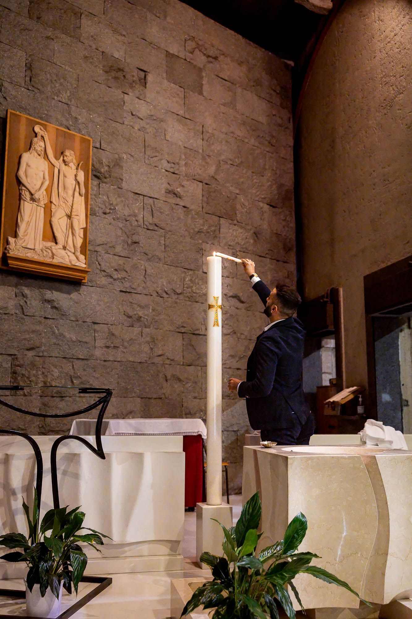 Man in suit lighting a tall white candle at a church altar with stone wall and religious relief artwork.