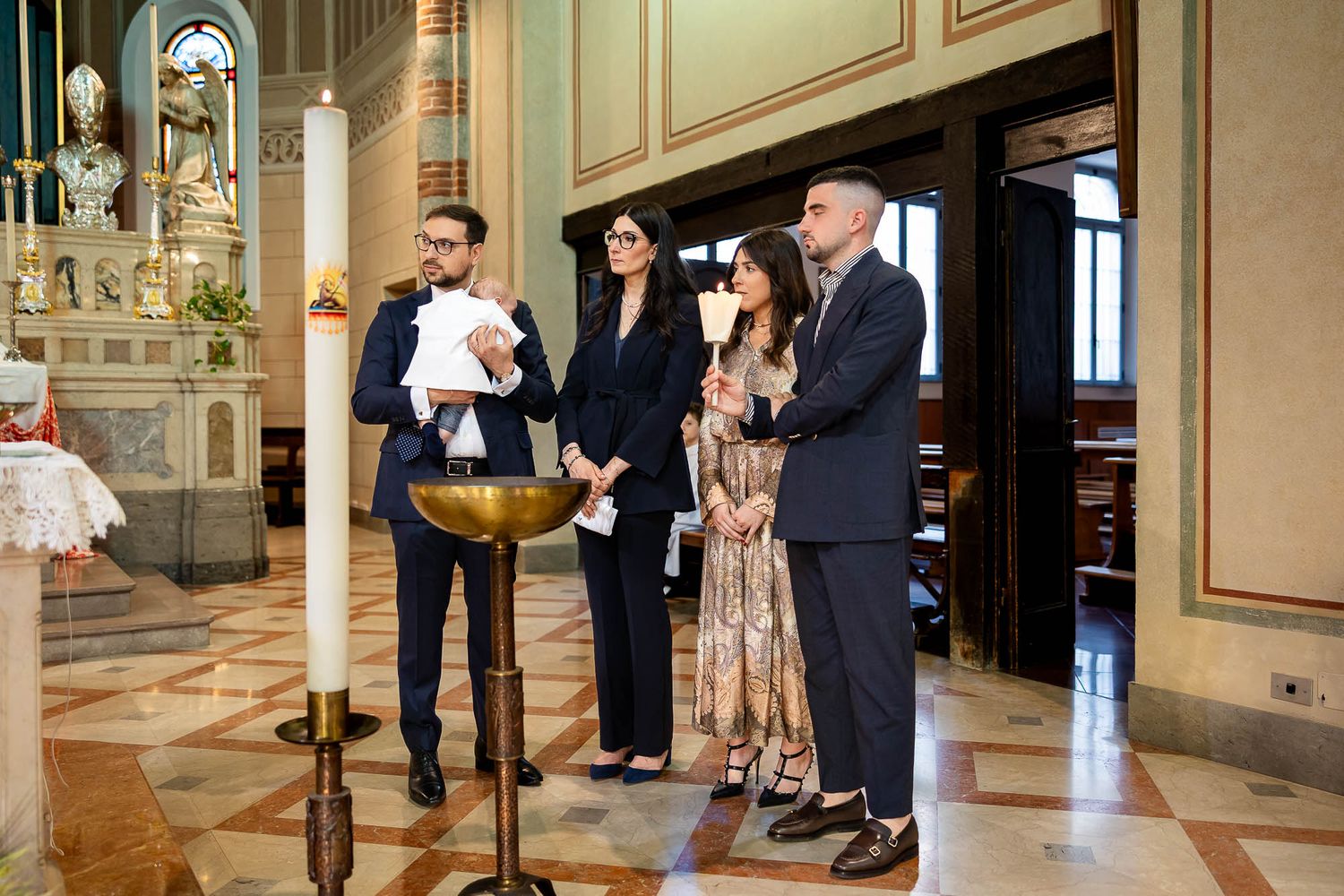 A baptism ceremony in a church with four adults standing near a baptismal font and lit candle.
