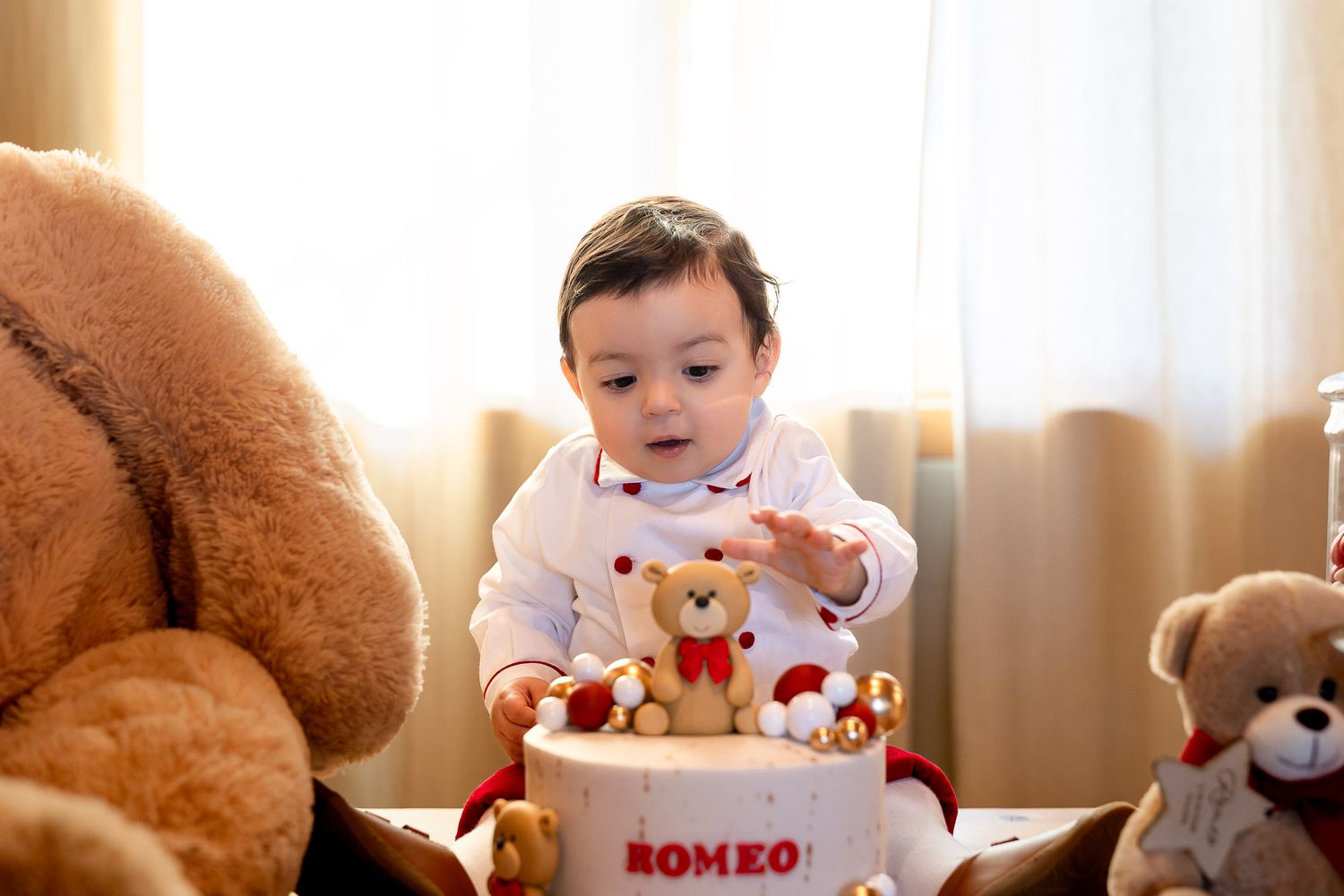 Smiling baby Romeo celebrates his first birthday with a teddy bear cake and stuffed animals nearby.