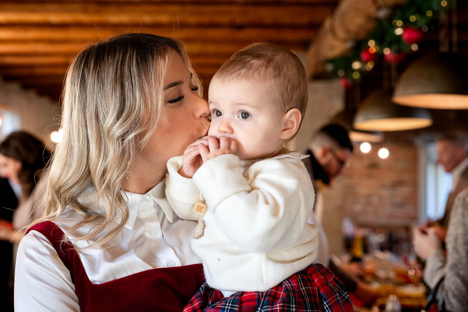 A blonde mother kisses her baby dressed in festive plaid at a Christmas family gathering.