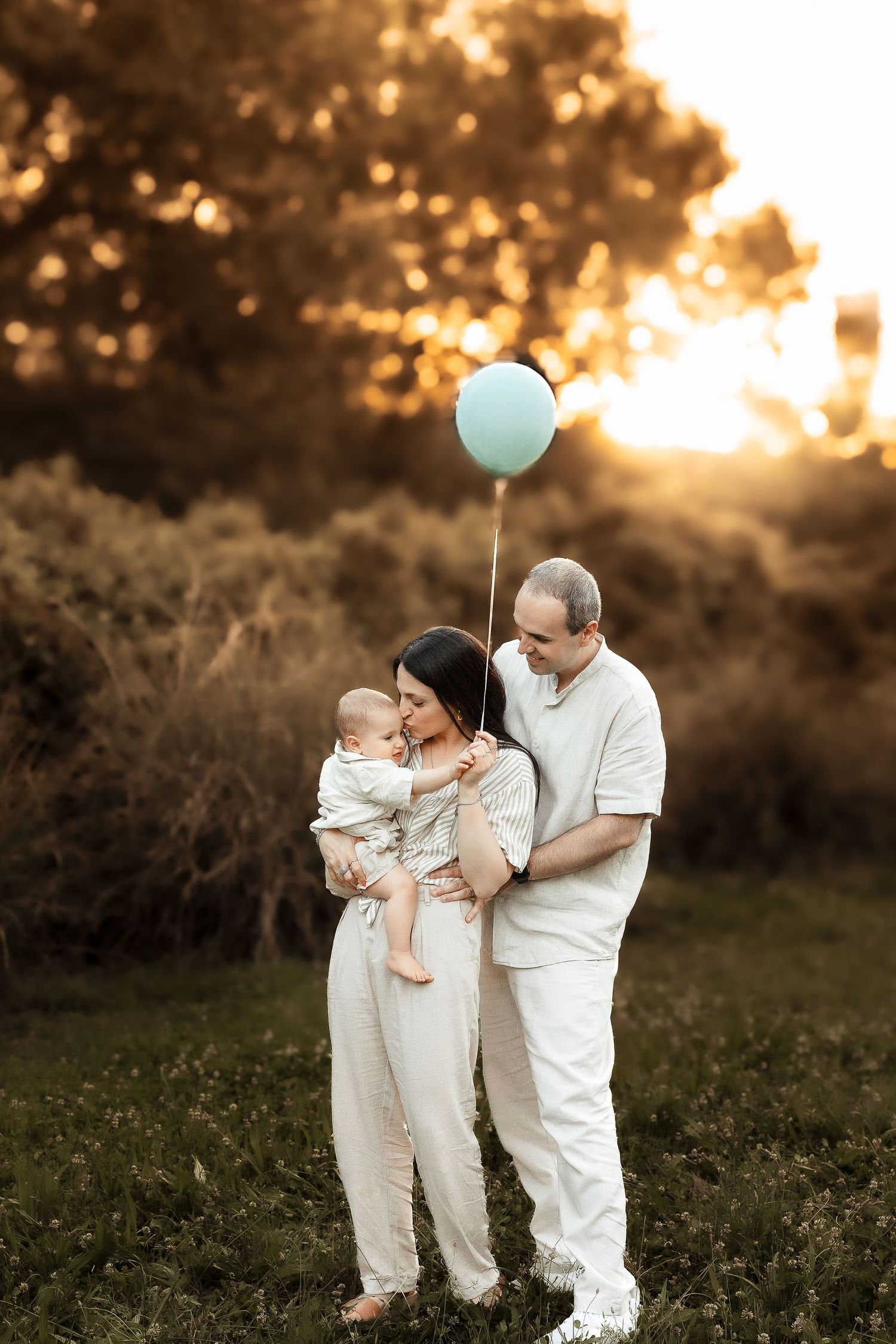 Family with baby holding teal balloon at golden sunset in park, wearing white outfits.