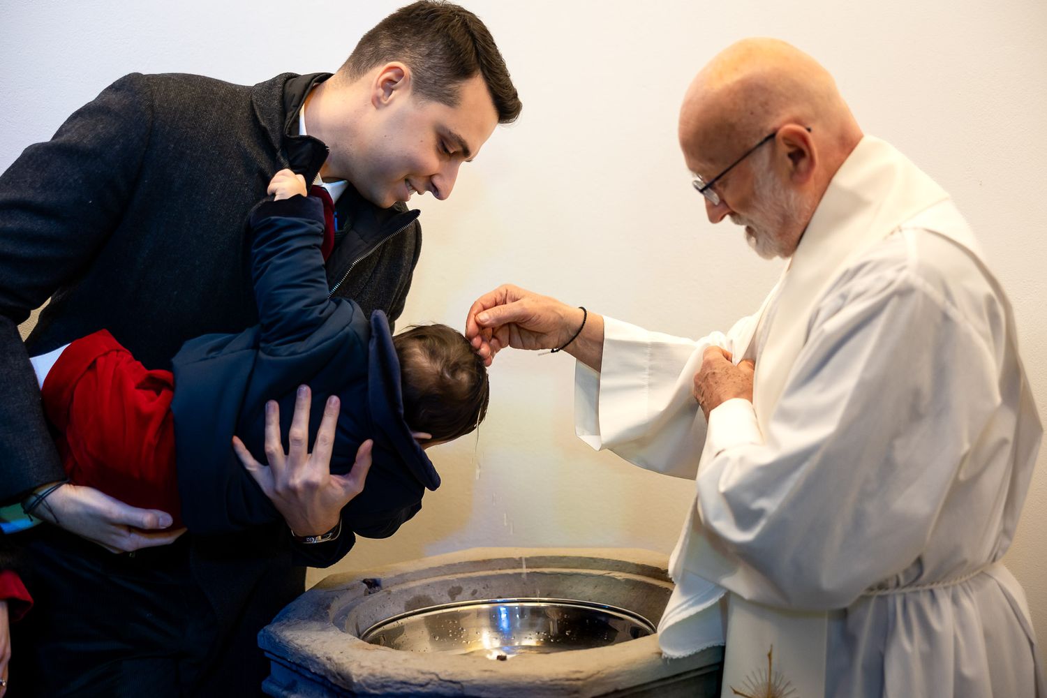 Priest performing a Catholic baptism ceremony, pouring holy water over infant held by parent at baptismal font.