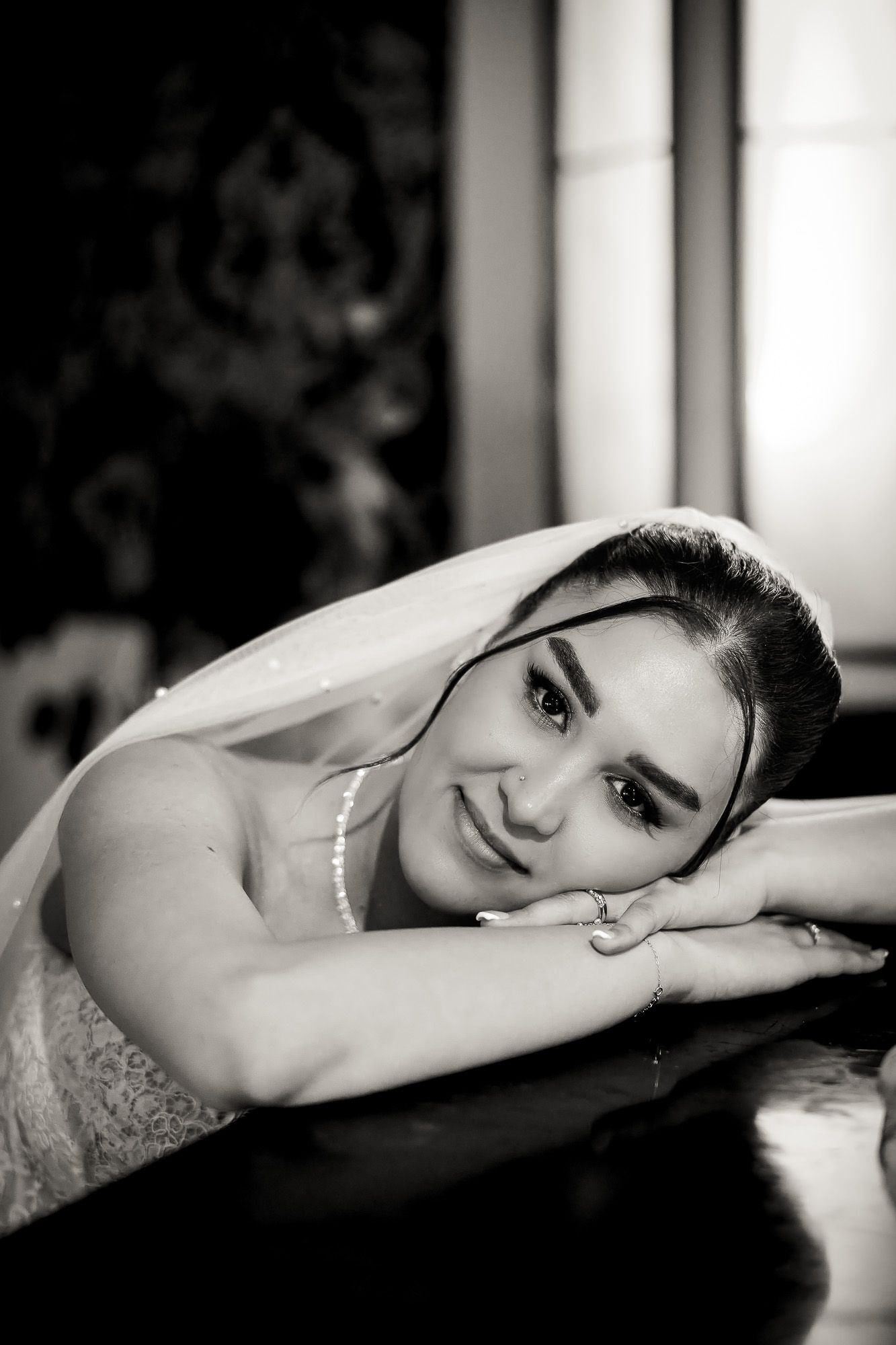Black and white portrait of a smiling bride in veil resting her chin on hands, elegant wedding photography.