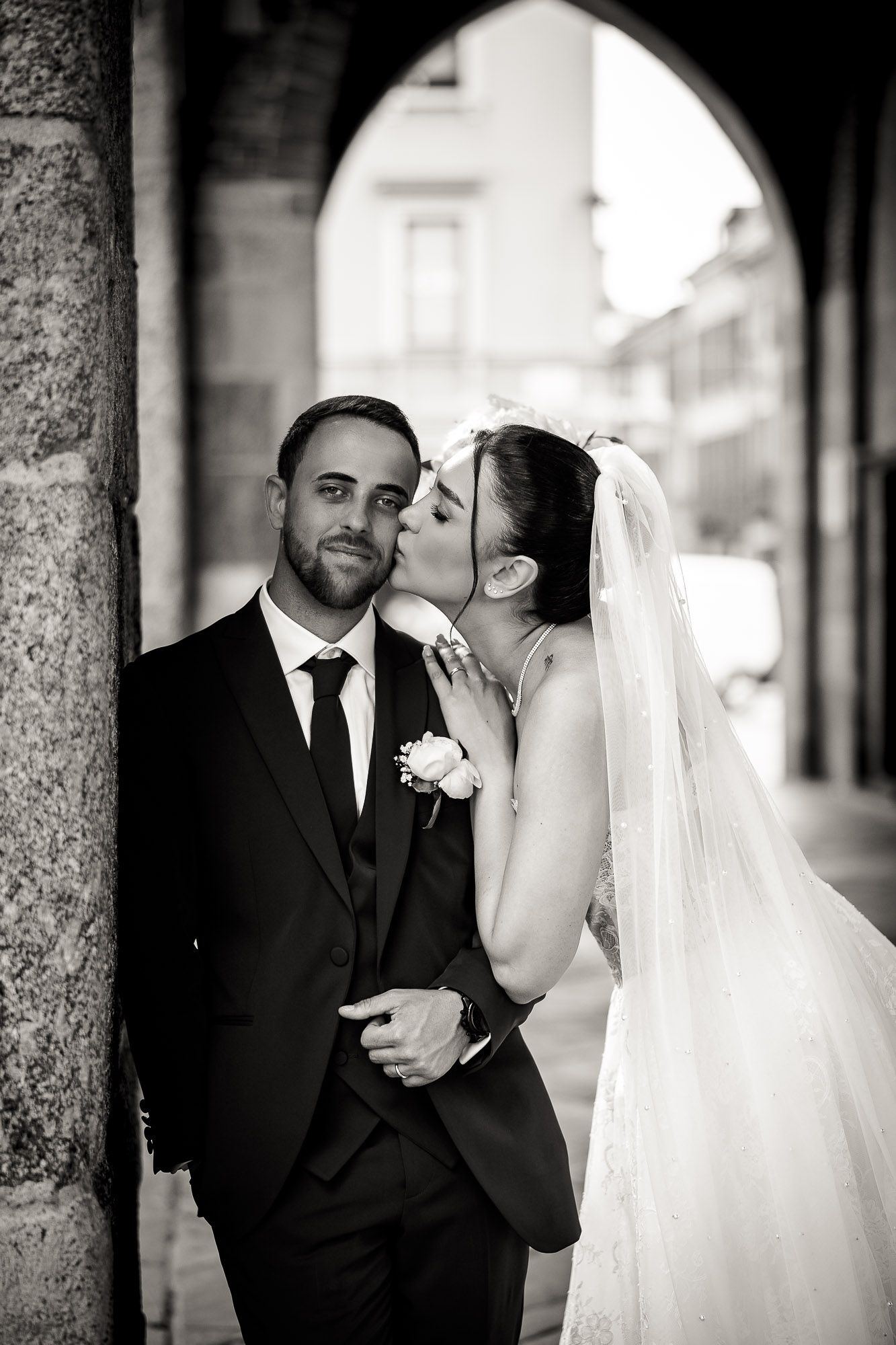 Black and white wedding photo of bride kissing groom on cheek under stone arch.