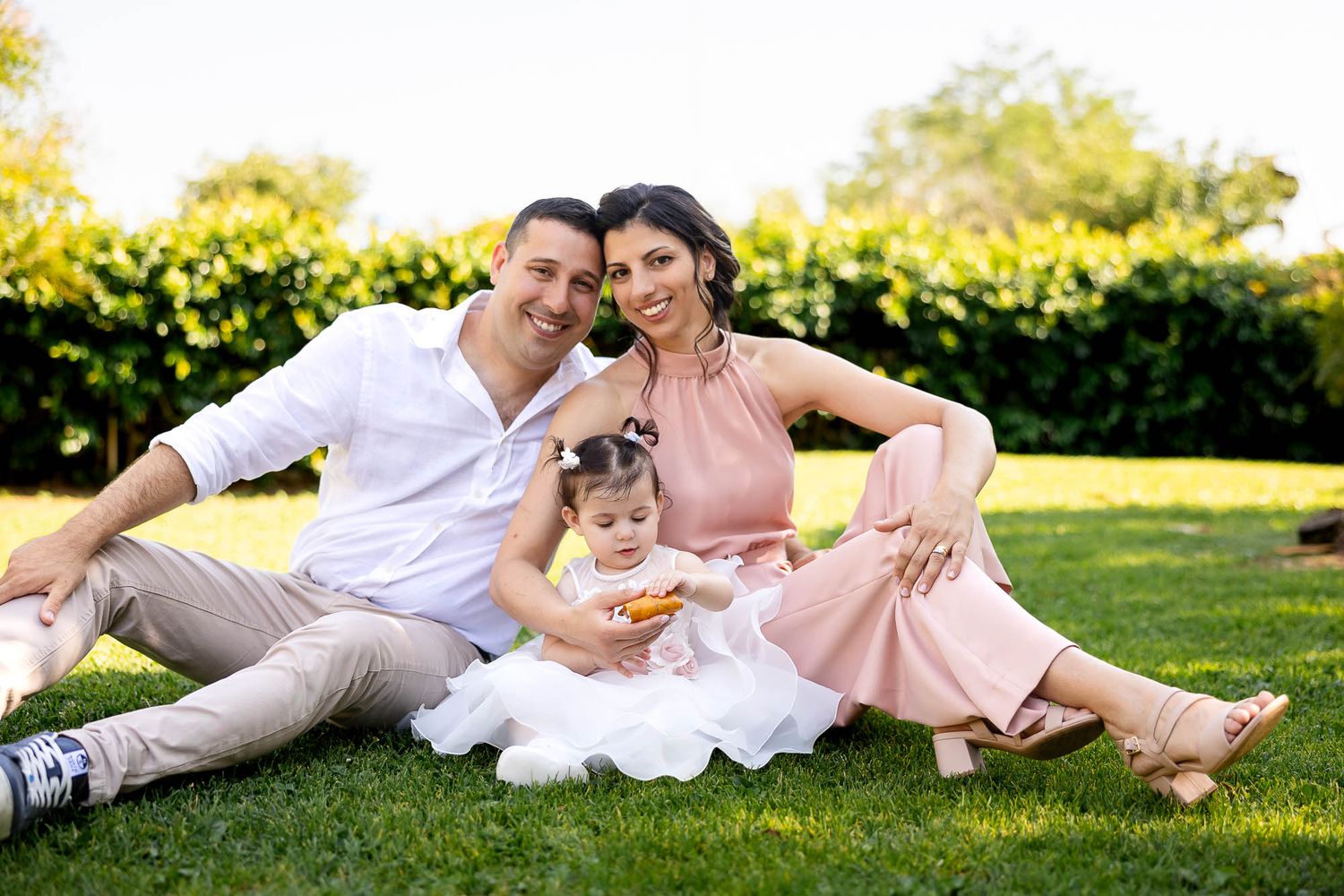 Family of three sitting on green grass in sunny park, parents in pink and white outfits with toddler girl between them.