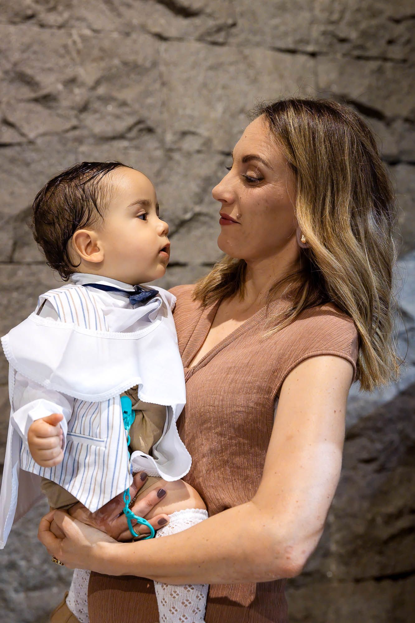 A young woman holds a dressed baby boy, both gazing at each other lovingly near a stone wall.