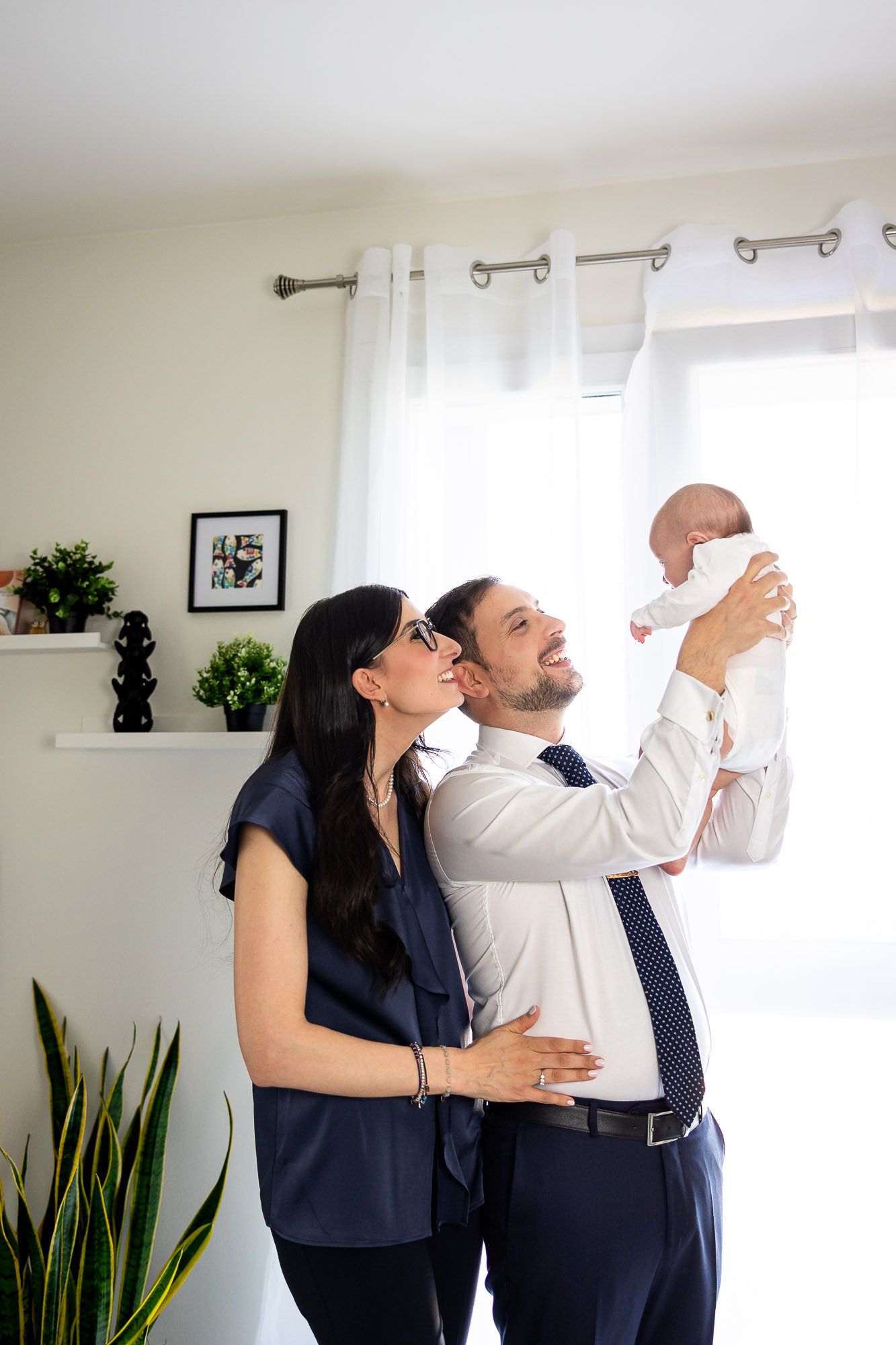 A happy couple in formal attire holds their newborn baby up in a bright, modern living room.