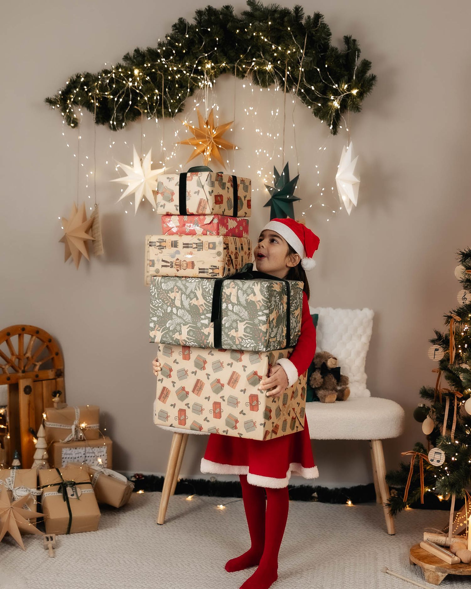 Child in Santa costume holding stack of wrapped Christmas gifts beside decorated tree with fairy lights.